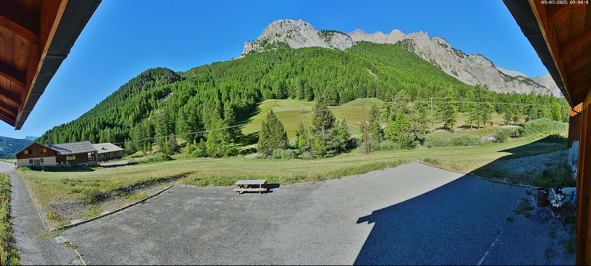 A gravel area with a picnic table sits in front of a building, overlooking a green valley and a large, mountainous backdrop under a clear blue sky.