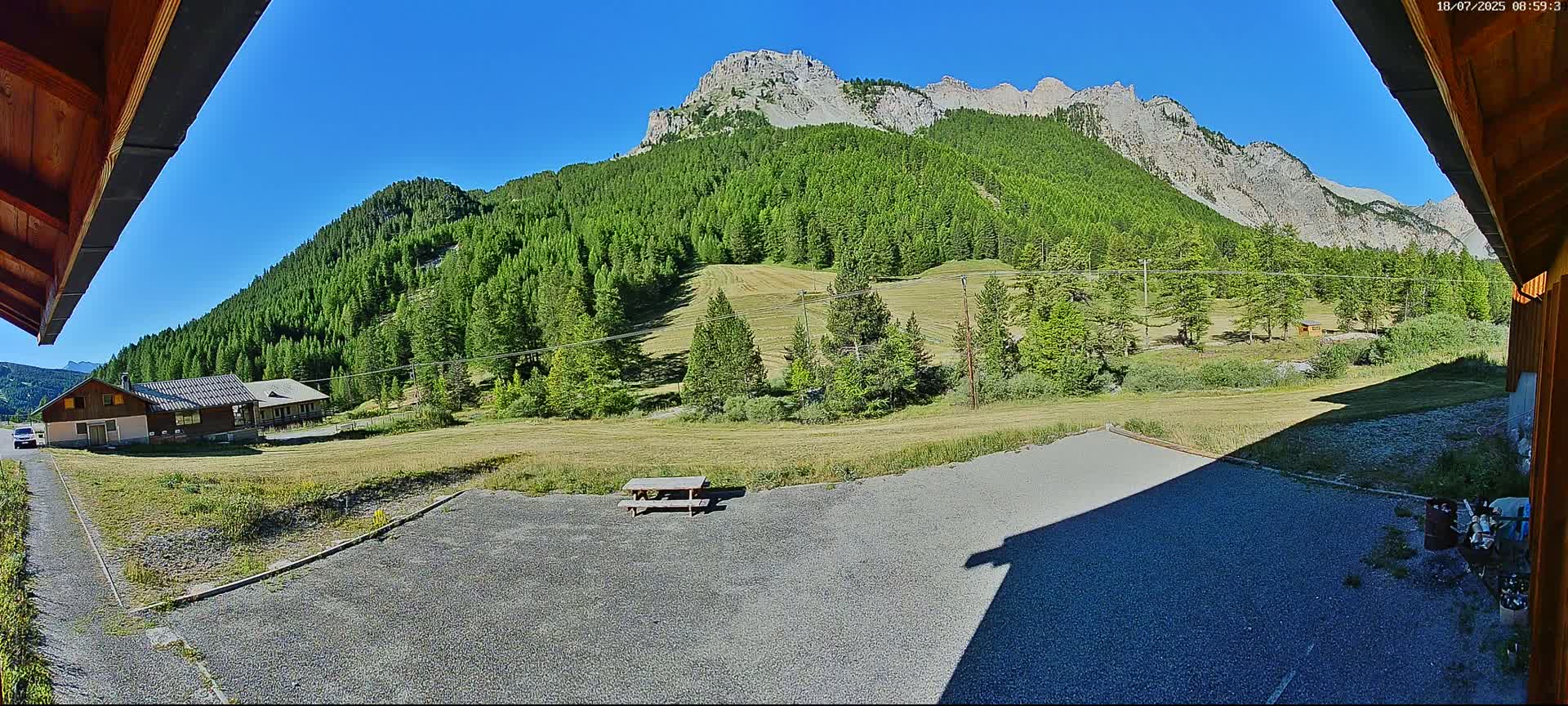 A gravel area with a picnic table sits in front of a building, overlooking a grassy field, a dense forest covering a mountainside, and a clear blue sky.