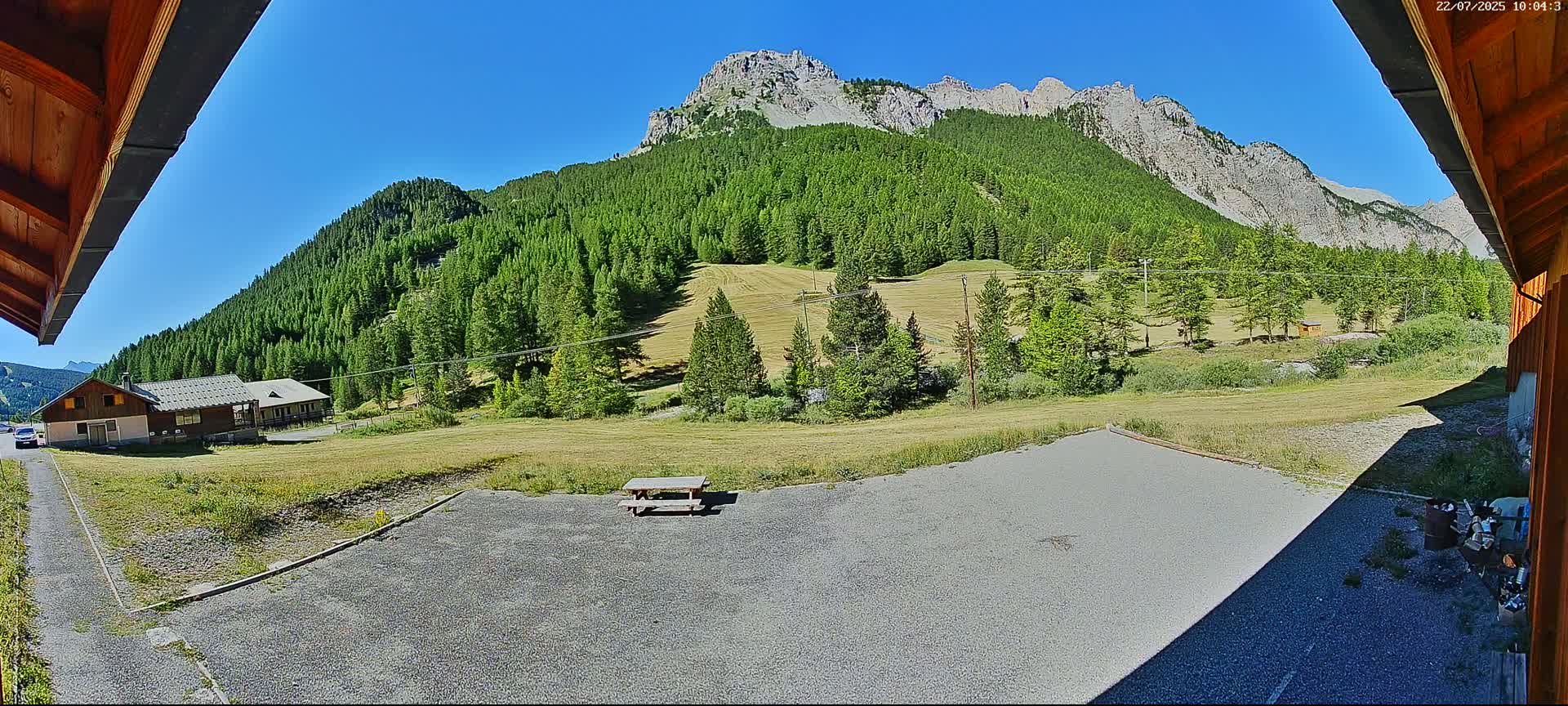 A sunny mountain vista is seen from a gravel parking area, featuring a building, picnic table, and a vast green hillside with a rocky peak in the background.