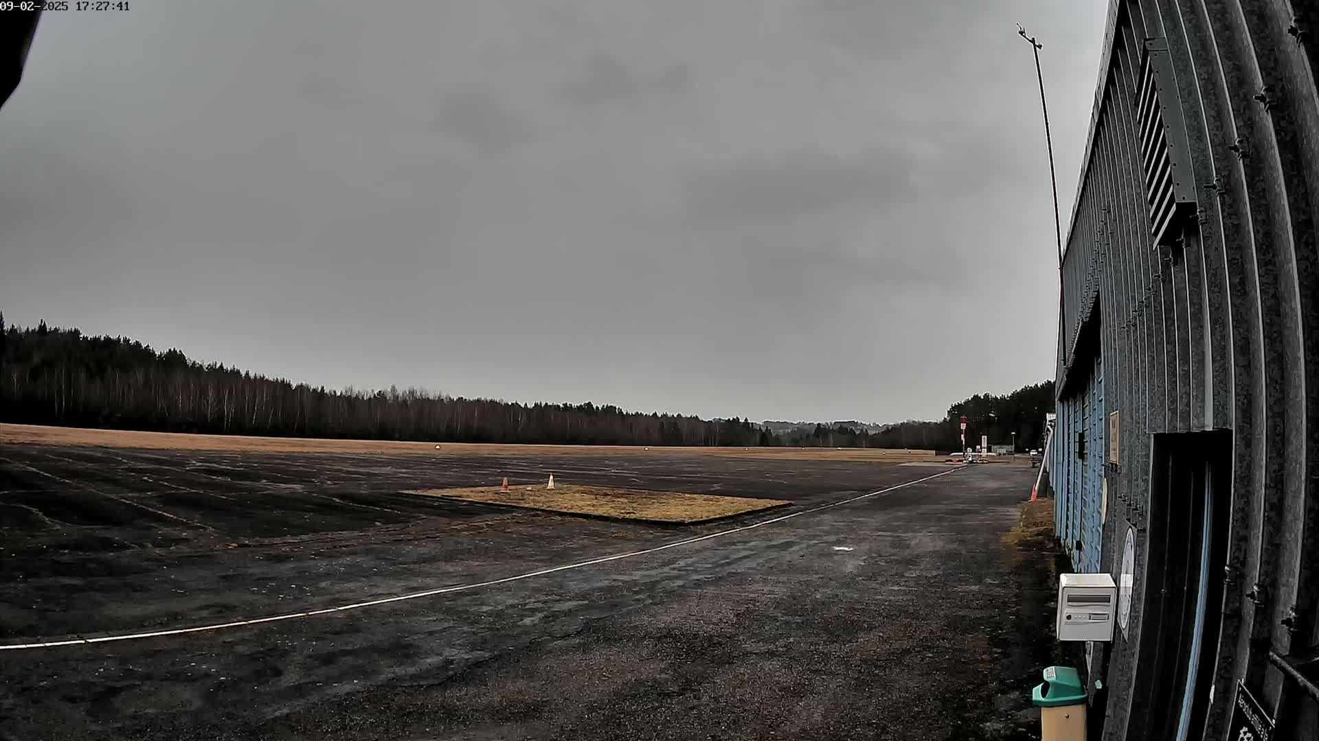 A dark gray overcast sky looms over a flat, dark tarmac area bordered by a line of trees and a corrugated metal building.