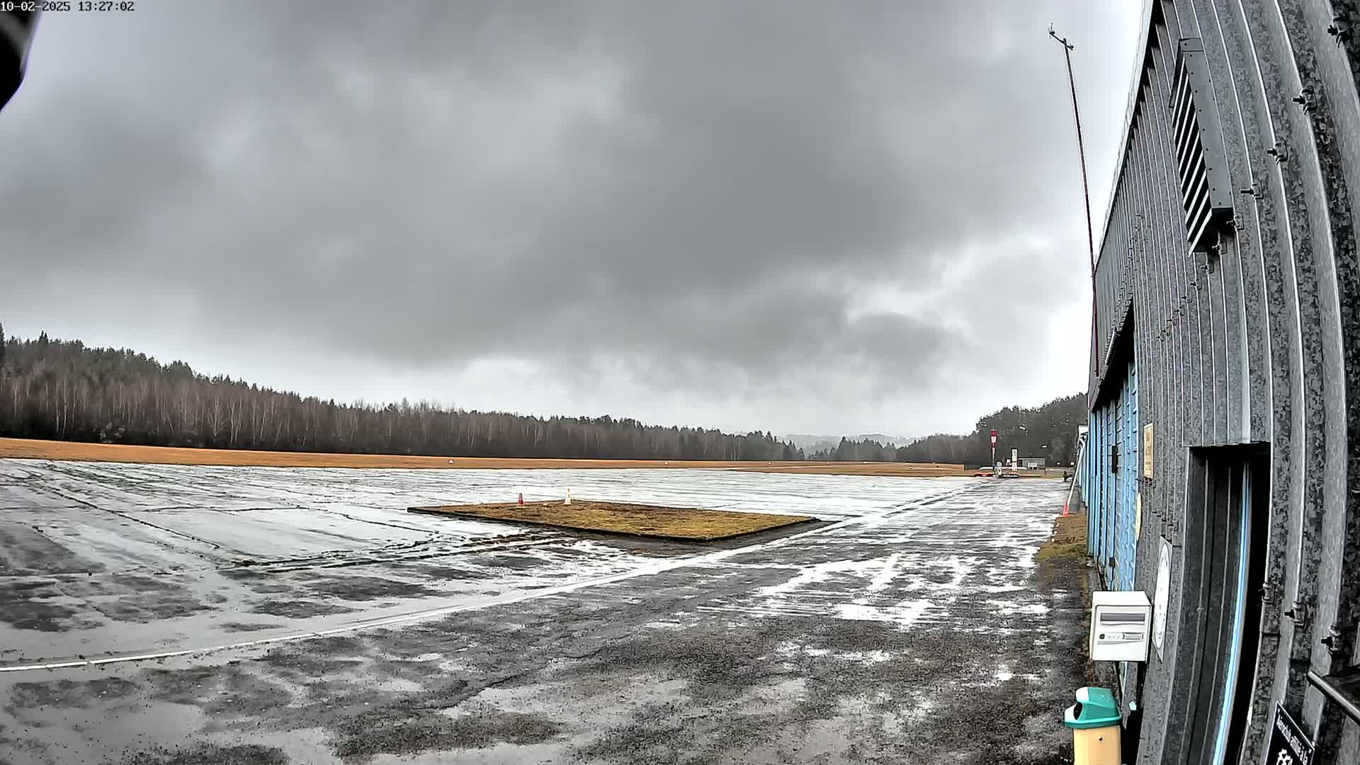 A wet, overcast airfield with a small grassy patch in the center, bordered by a line of trees and a hangar to the right.
