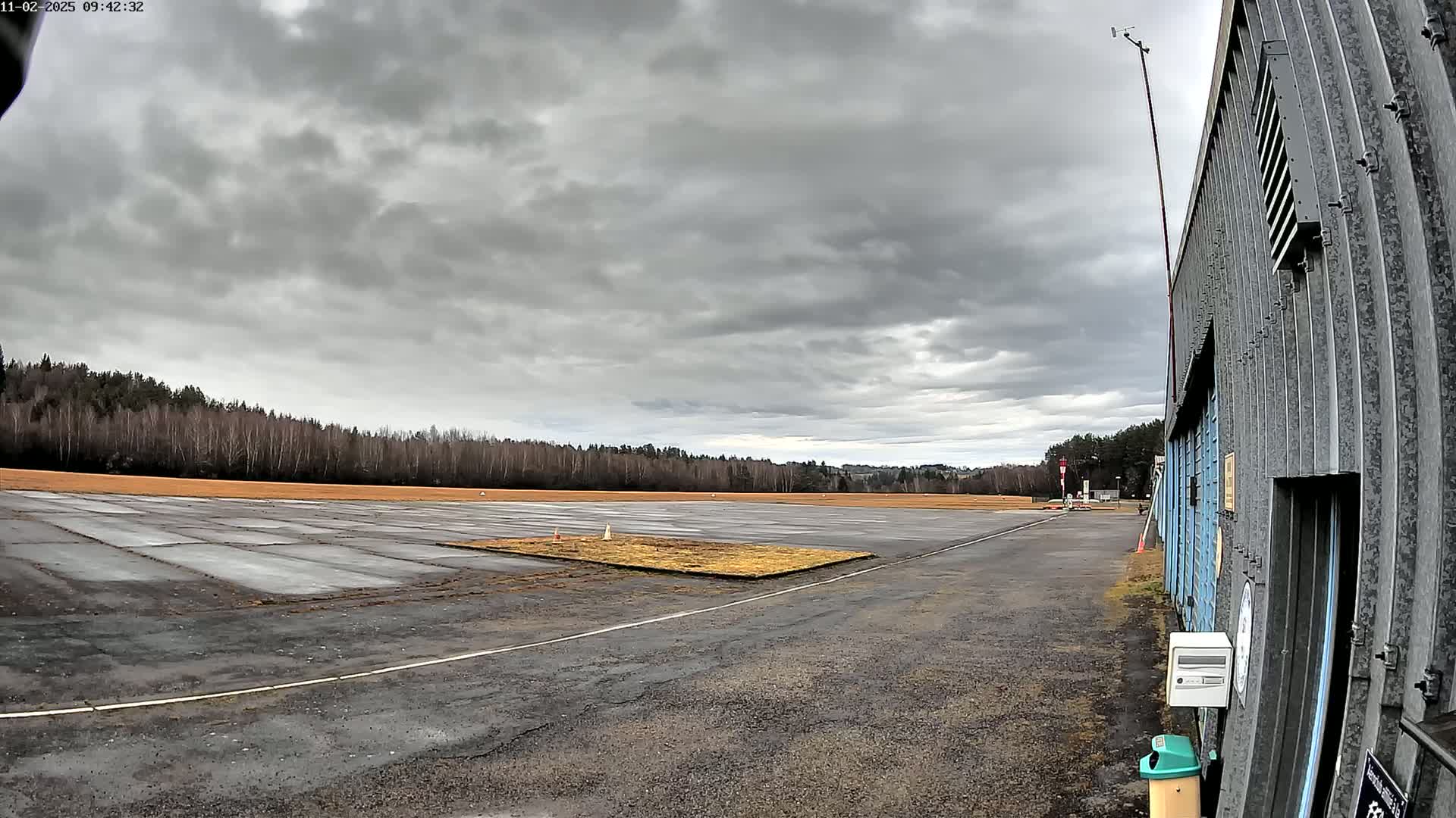 A mostly overcast sky hangs over a flat, paved area next to a hangar, bordered by a line of leafless trees.