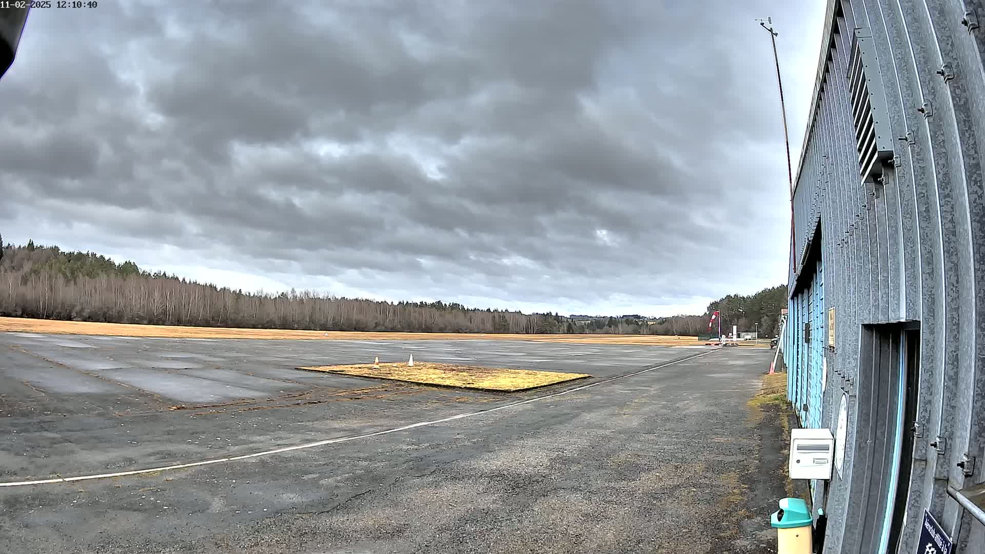 A gray, overcast sky hangs over a flat, paved airfield bordered by a line of bare trees and a corrugated metal hangar.