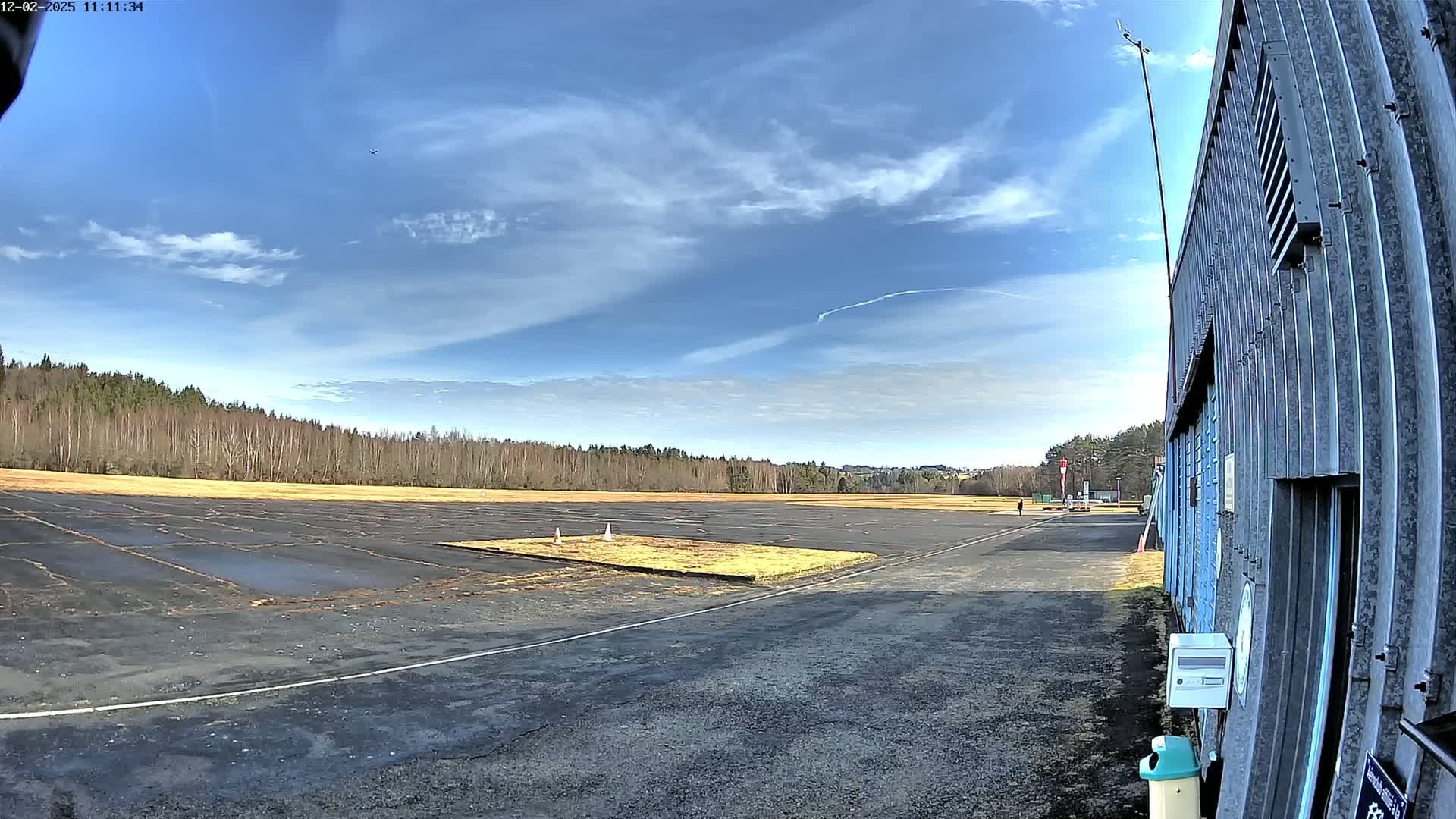 A paved airfield with a grassy area in the center stretches to a line of bare trees under a mostly sunny sky.