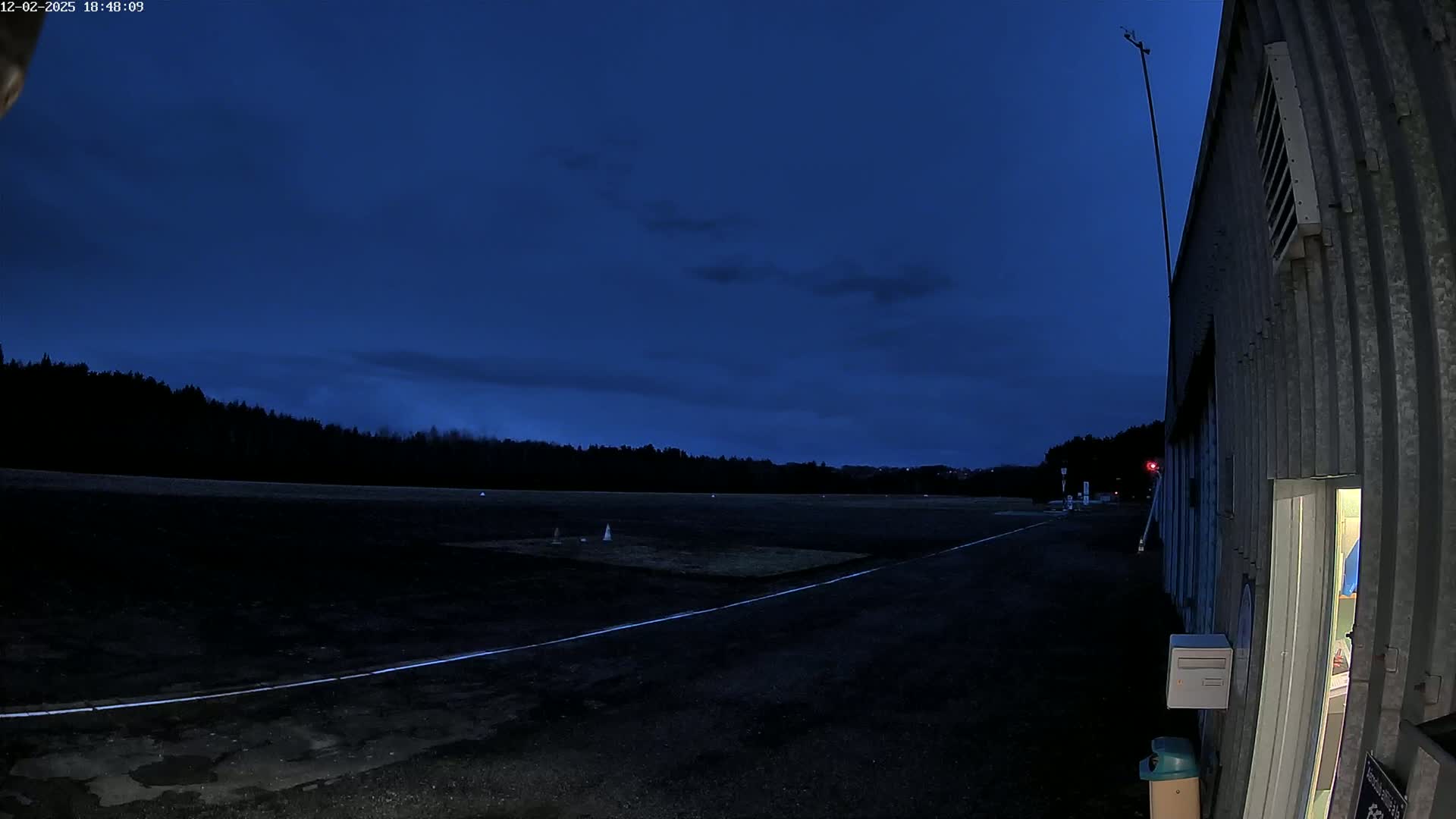 A dark asphalt area with white lines, possibly a runway or airfield, is adjacent to a corrugated metal building under a twilight sky.