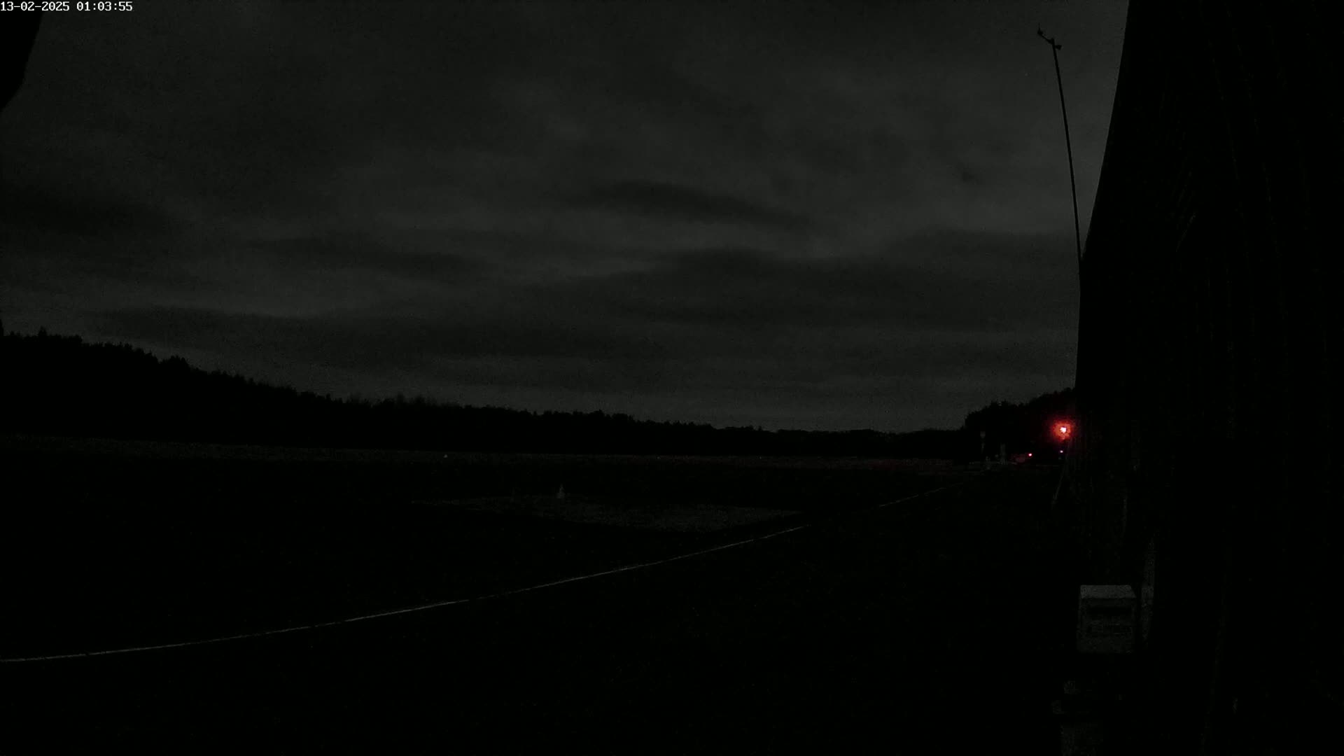 A dark, nighttime scene shows a flat, dark landscape under a mostly cloudy sky, with a single faint red light visible in the distance.