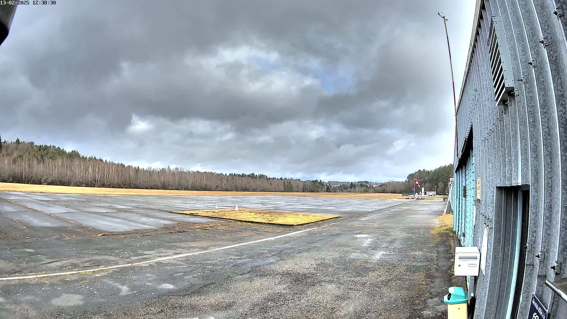 A wet, paved airstrip extends to a line of bare trees under a cloudy sky, with a corrugated metal building visible at the right.