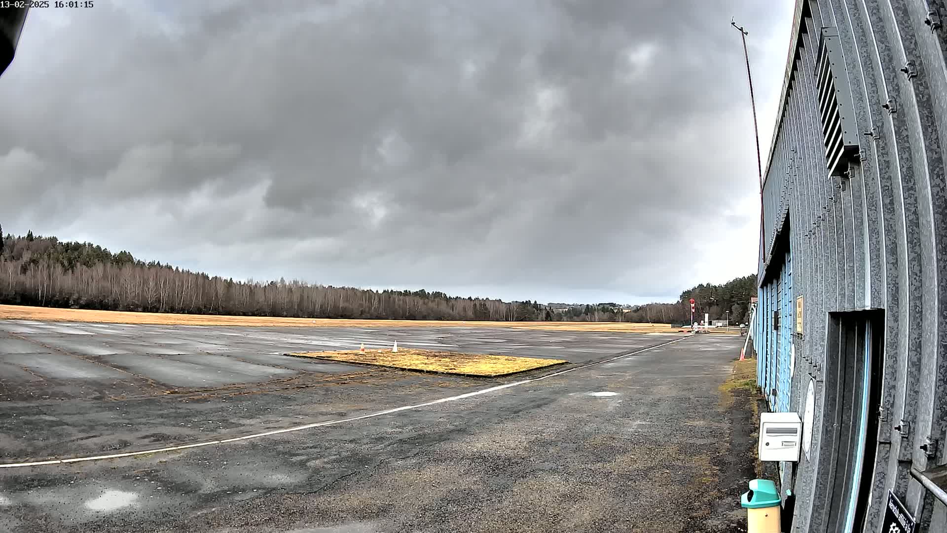 A mostly overcast sky hangs over a tarmac airfield bordered by a line of trees and a corrugated metal hangar.