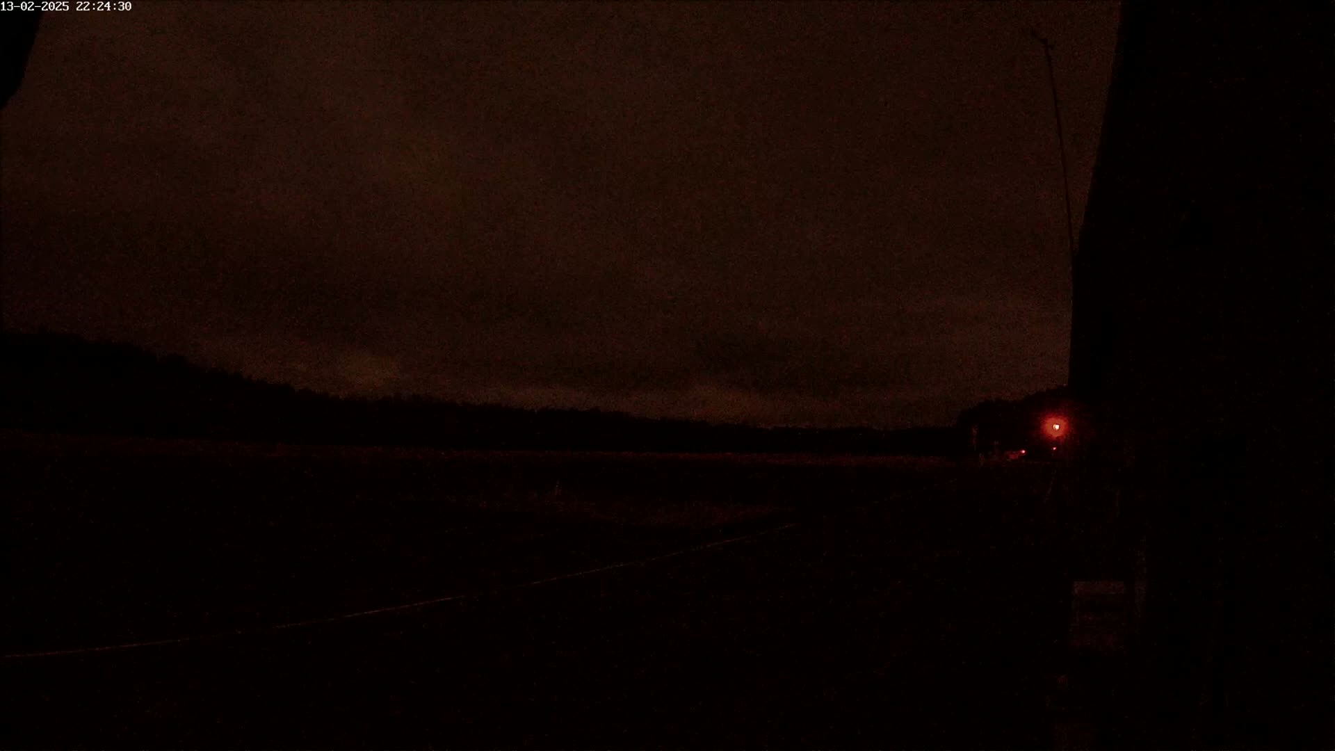 A dark, nighttime scene shows a flat, dark landscape under a mostly cloudy sky, with a single faint red light visible in the distance.