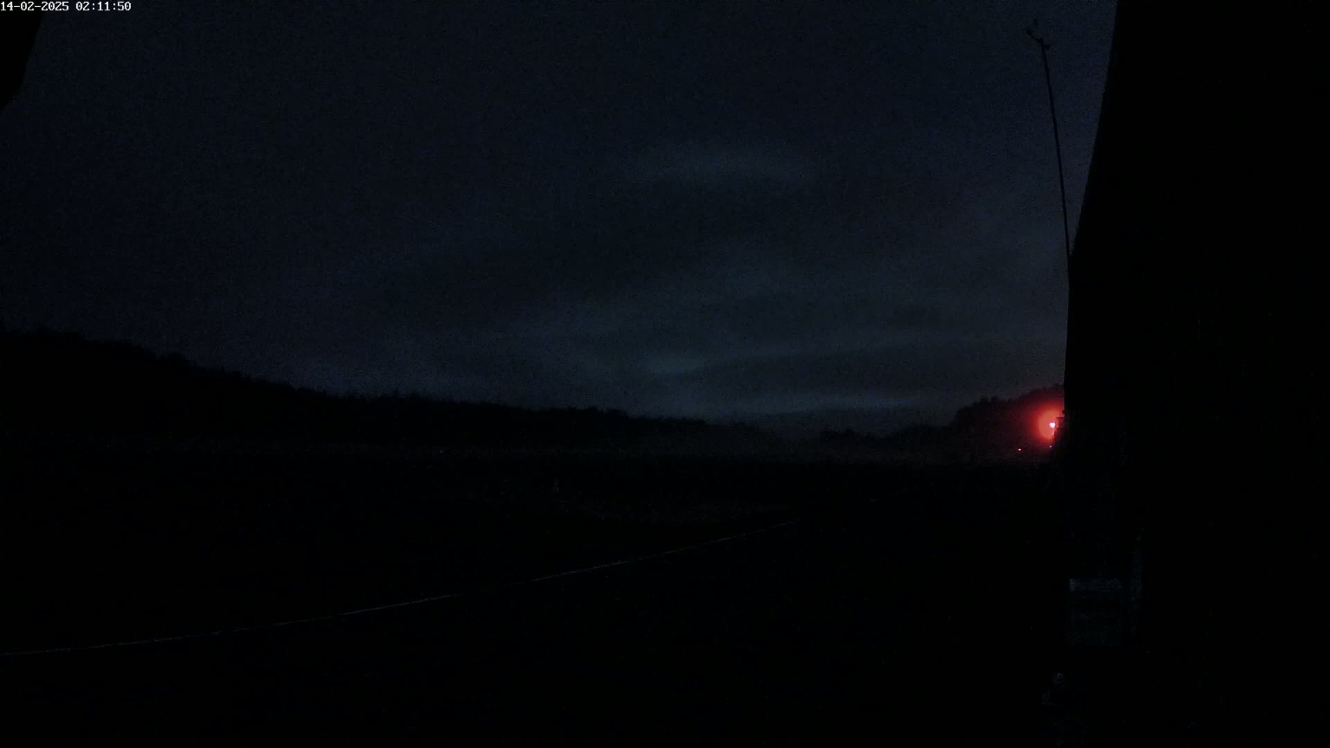 A dark, nighttime scene shows a distant, dimly lit landscape under a cloudy sky, partially obscured by a large, dark structure in the foreground.