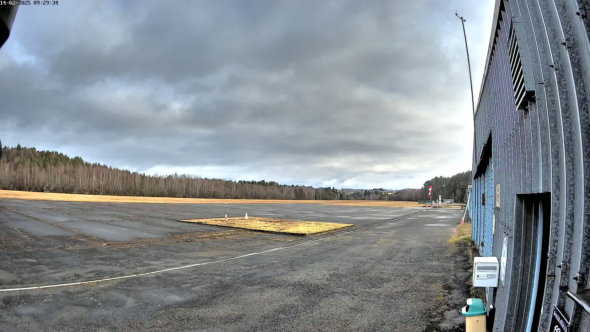 A mostly overcast sky hangs over a deserted airfield with a small grassy area in the center, adjacent to a metal building.