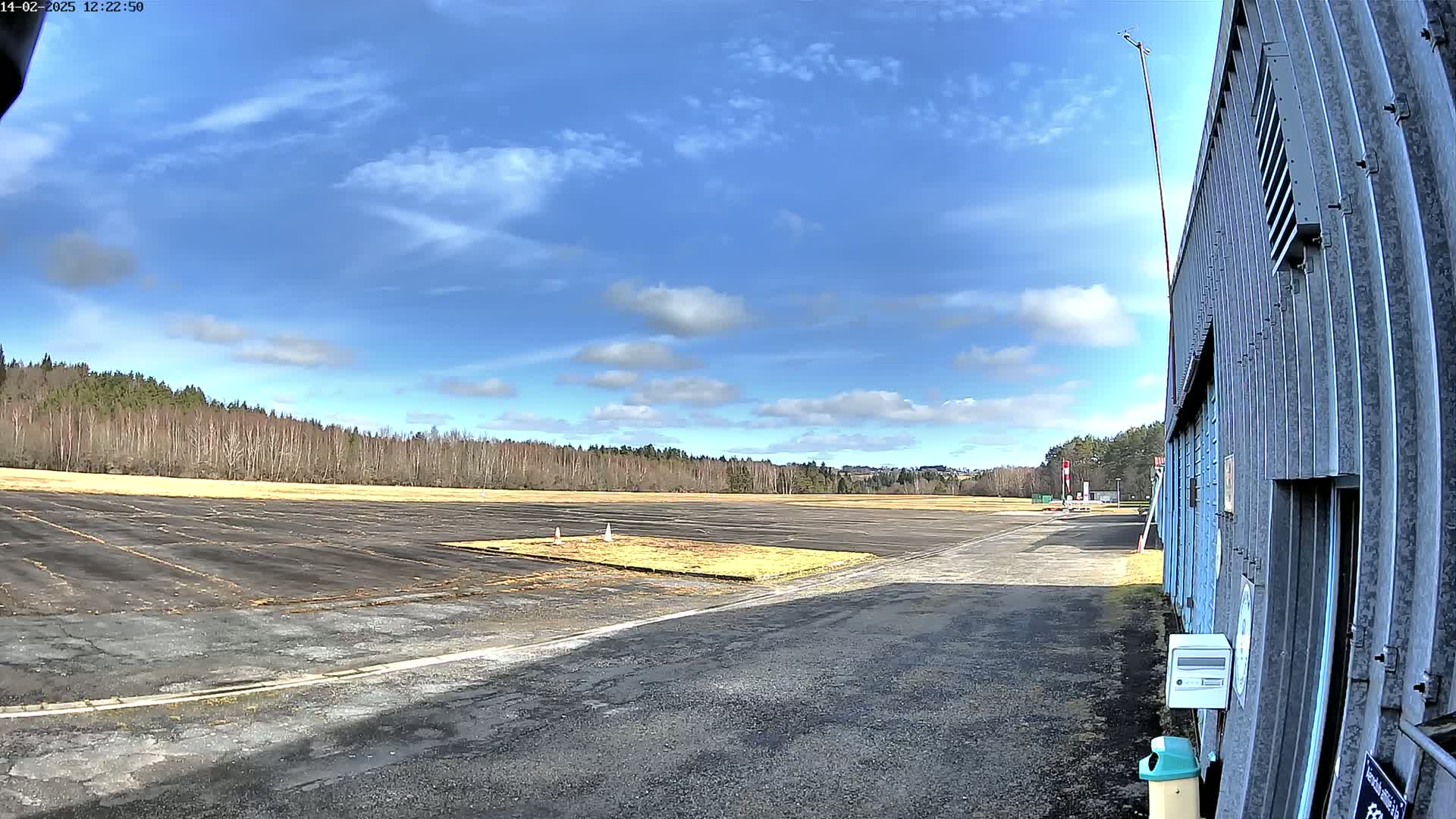 A mostly clear blue sky with scattered clouds overlooks a paved airfield bordered by a line of trees, next to a hangar.