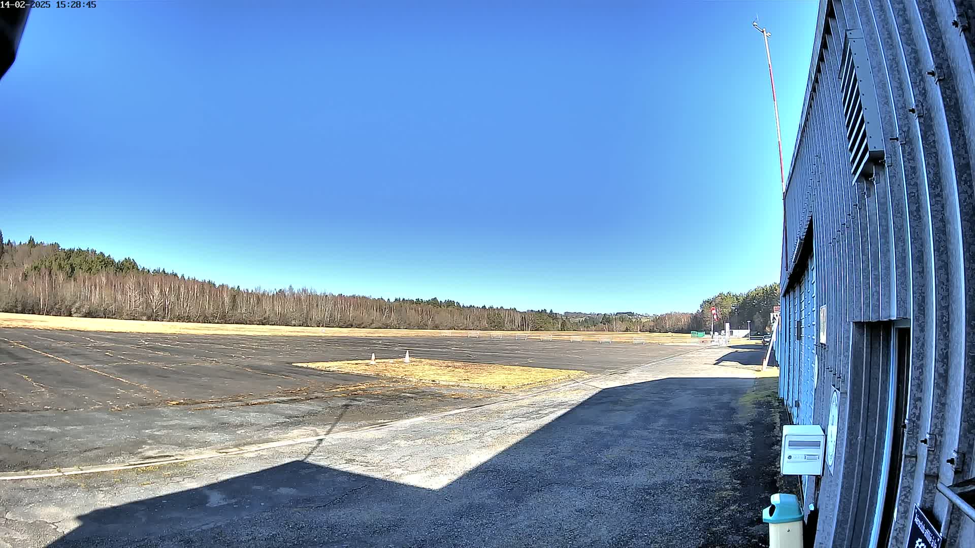 A large, empty paved area, possibly a runway or airfield, stretches out before a metal building under a clear blue sky.