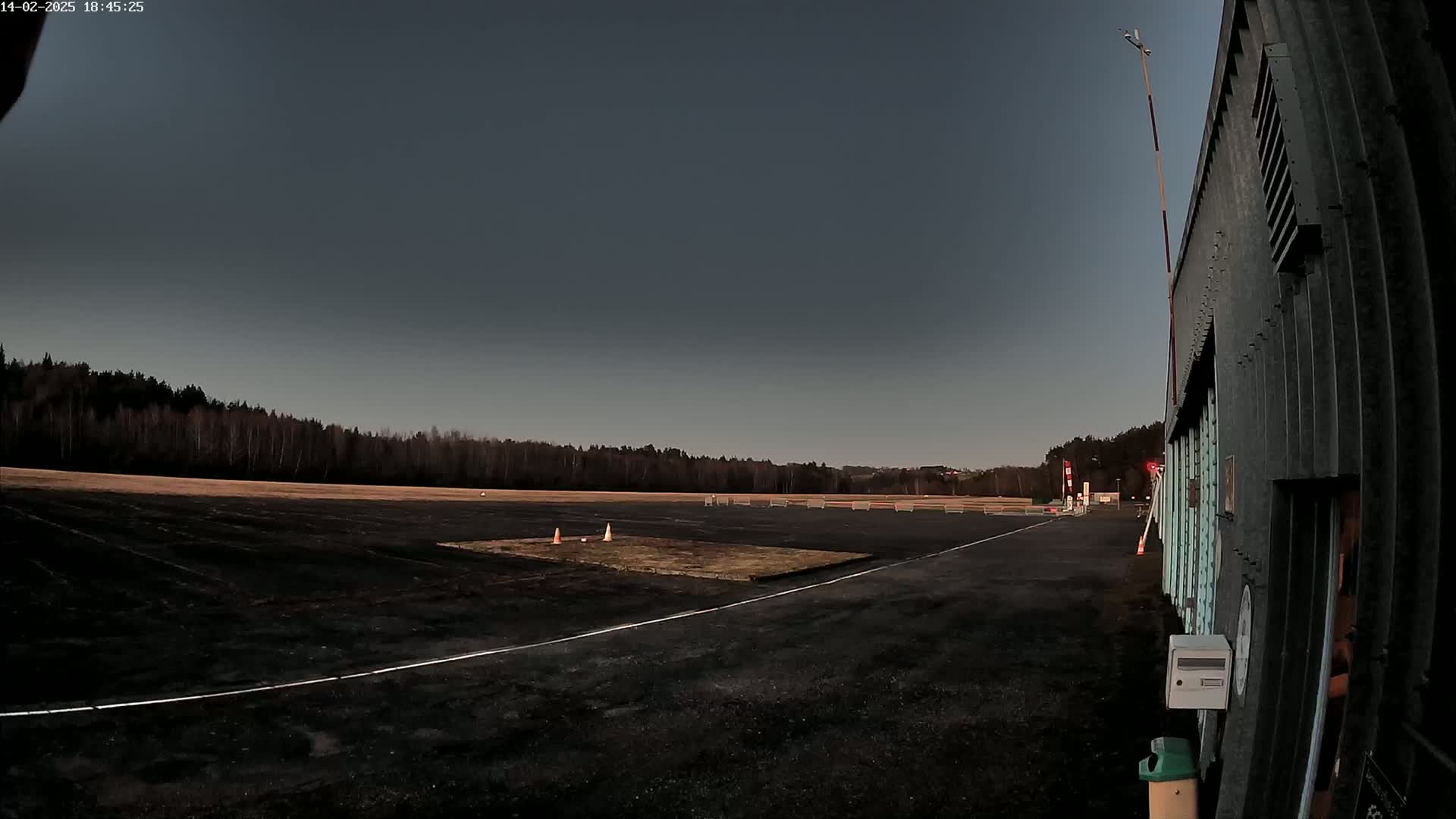 A dark asphalt area with white lines, a small patch of dirt, and traffic cones sits before a line of trees under a dusky sky.
