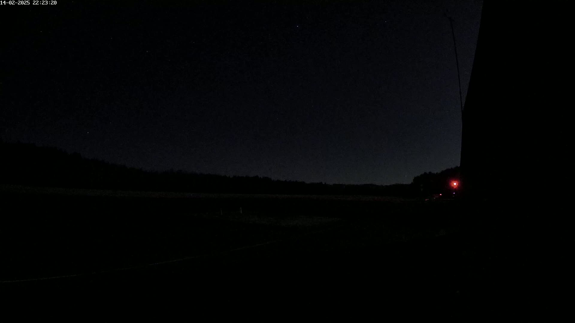 A dark, nighttime landscape featuring a distant treeline under a clear, starlit sky, with a single faint red light visible near the bottom right.