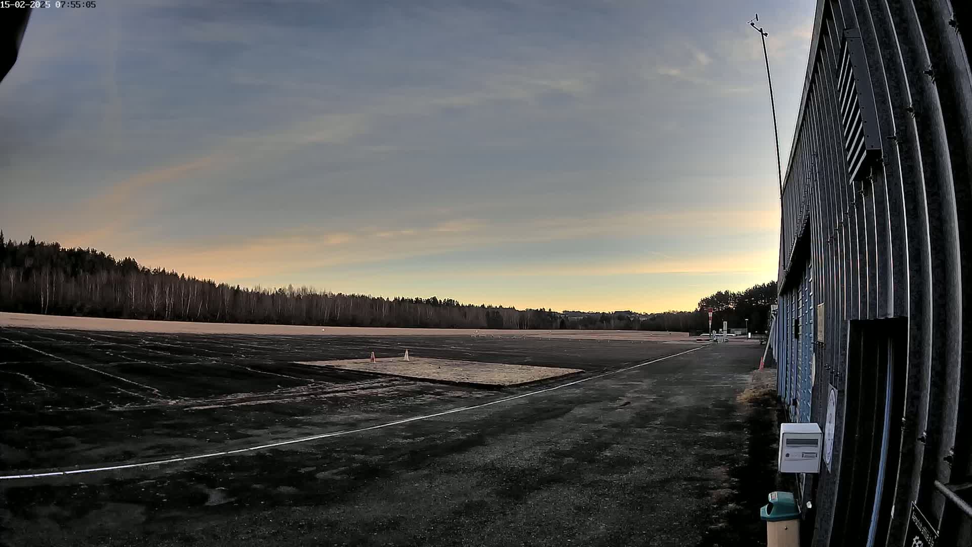 A dark, paved area with a smaller, lighter-colored patch in the center stretches towards a line of trees under a mostly clear, dusky sky.