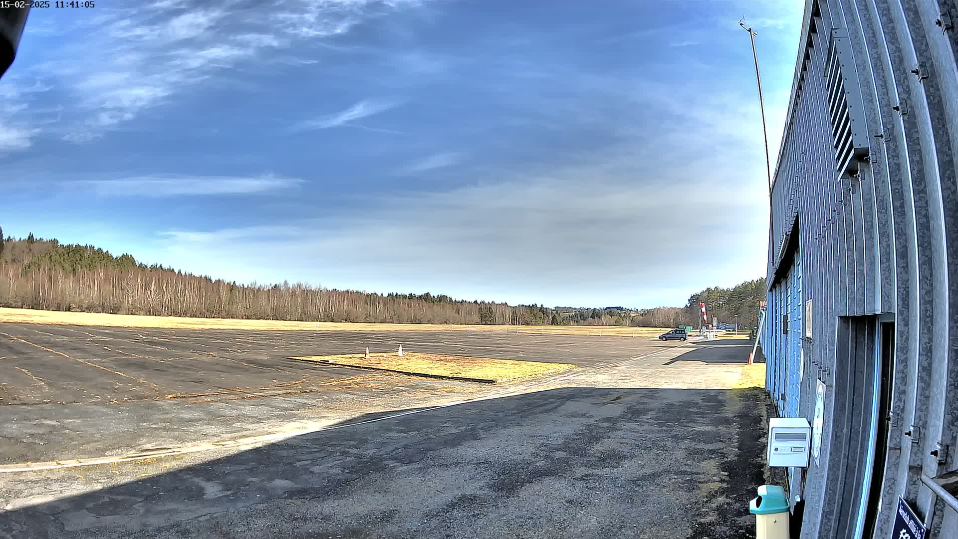 A large, mostly empty paved area, possibly a runway or airfield, is bordered by a line of trees under a mostly sunny sky with some scattered clouds, and a corrugated metal building is visible to the right.