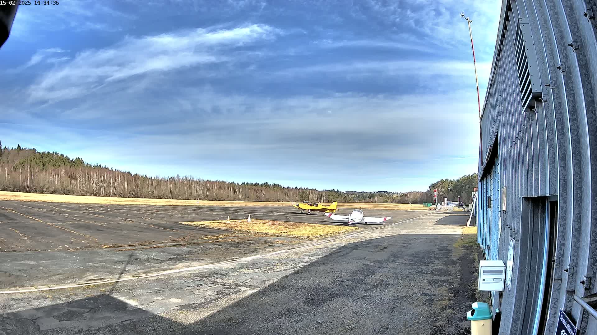 Two small airplanes are parked on a tarmac next to a hangar under a partly cloudy blue sky.