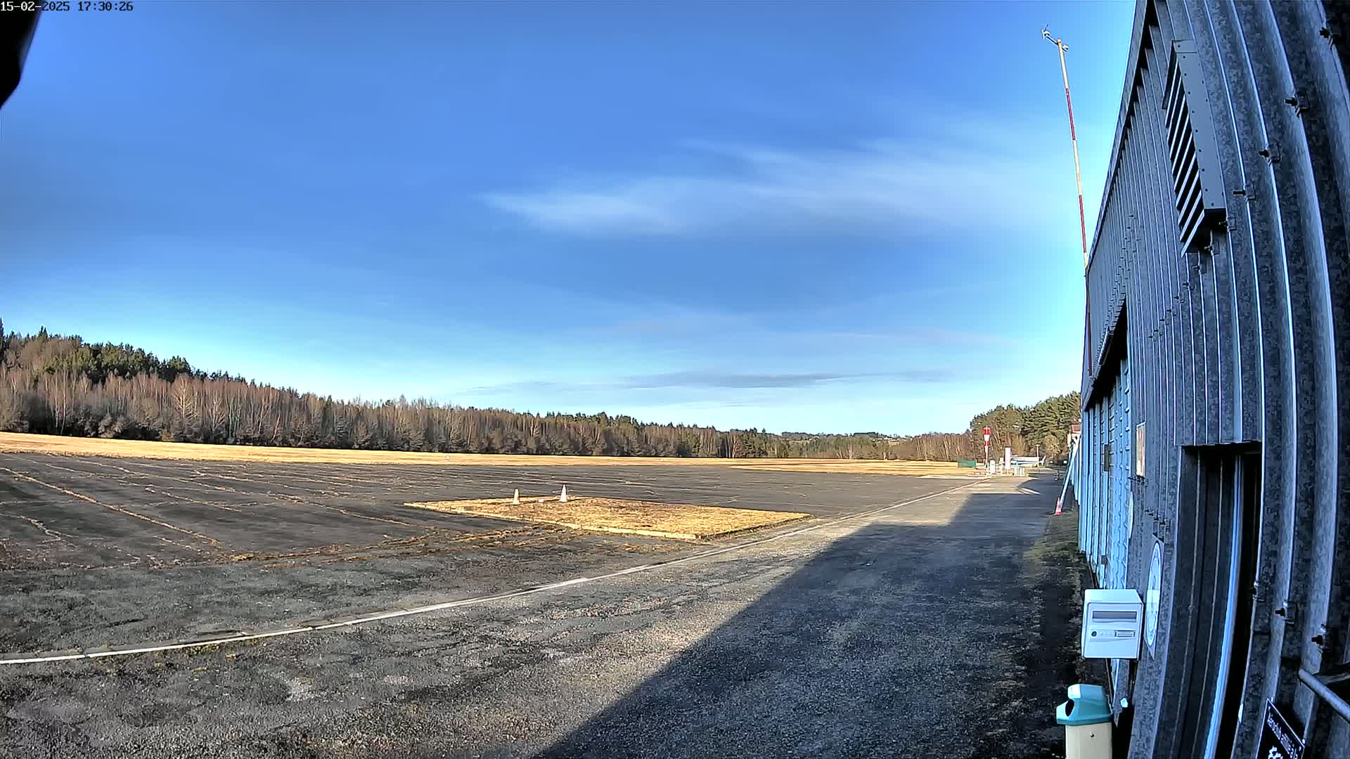 A mostly sunny day at a small airfield with a paved runway, bordered by a line of bare trees, and a corrugated metal building to the right.