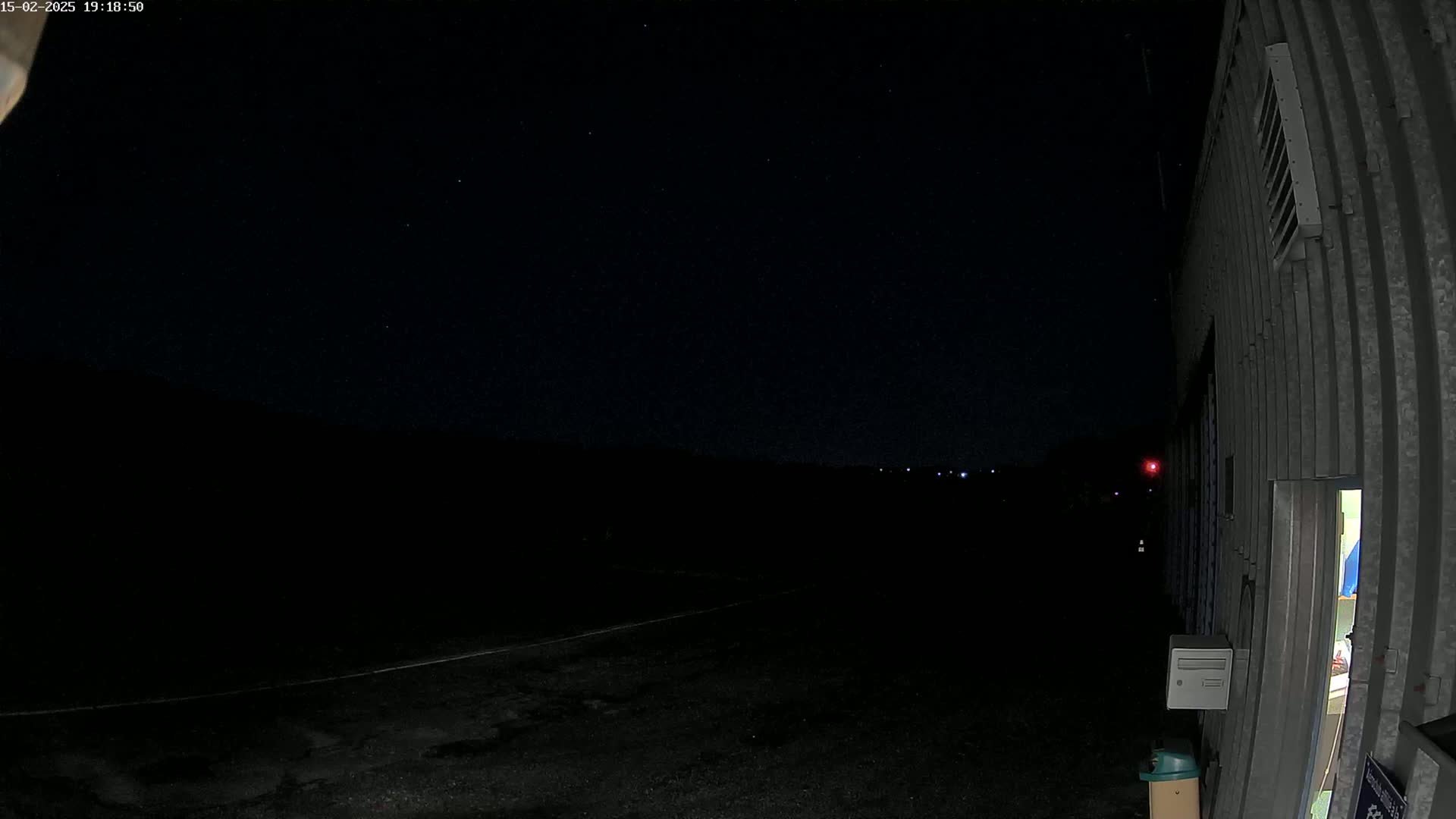 A dark, nighttime view of a paved area next to a corrugated metal building under a clear, starry sky.