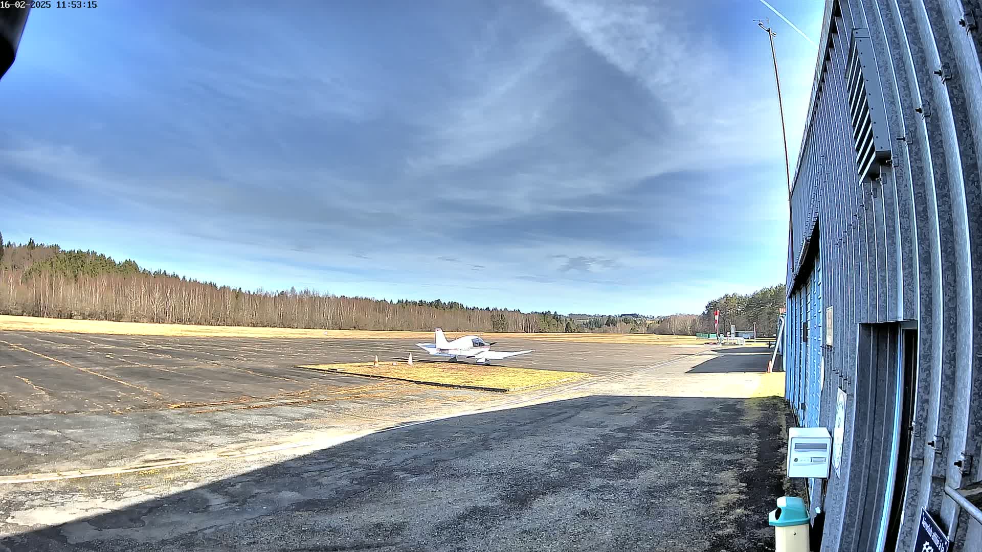 A small, white airplane sits on a tarmac in front of a hangar, with a line of trees and a partly cloudy blue sky in the background.