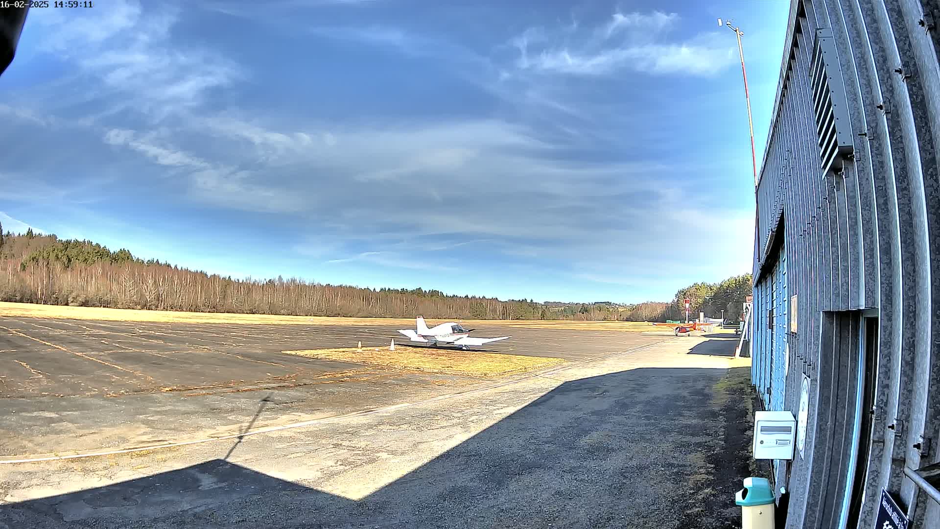 A small airplane sits on a tarmac next to a hangar, with a partly sunny sky and a line of trees in the background.