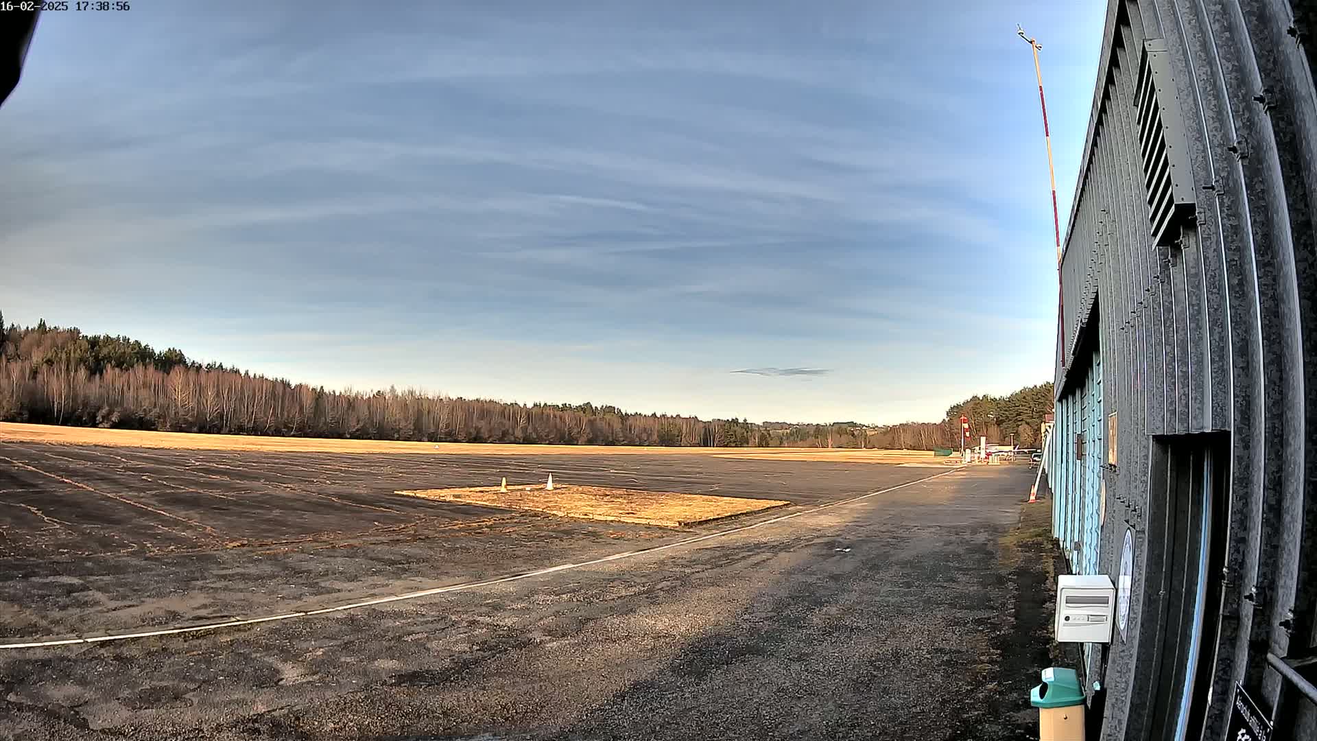 A mostly clear, late afternoon sky overlooks a paved airfield bordered by bare trees, with a hangar visible to the right.
