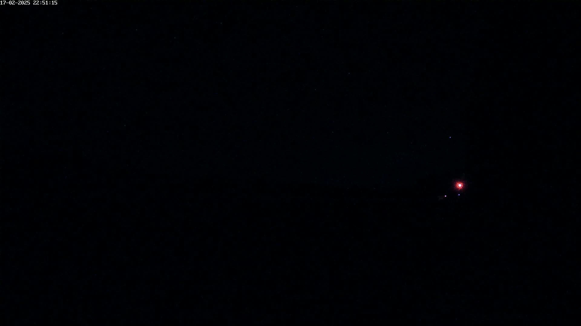A dark nighttime scene shows a small, faint, reddish light in the distance against a black sky speckled with stars.