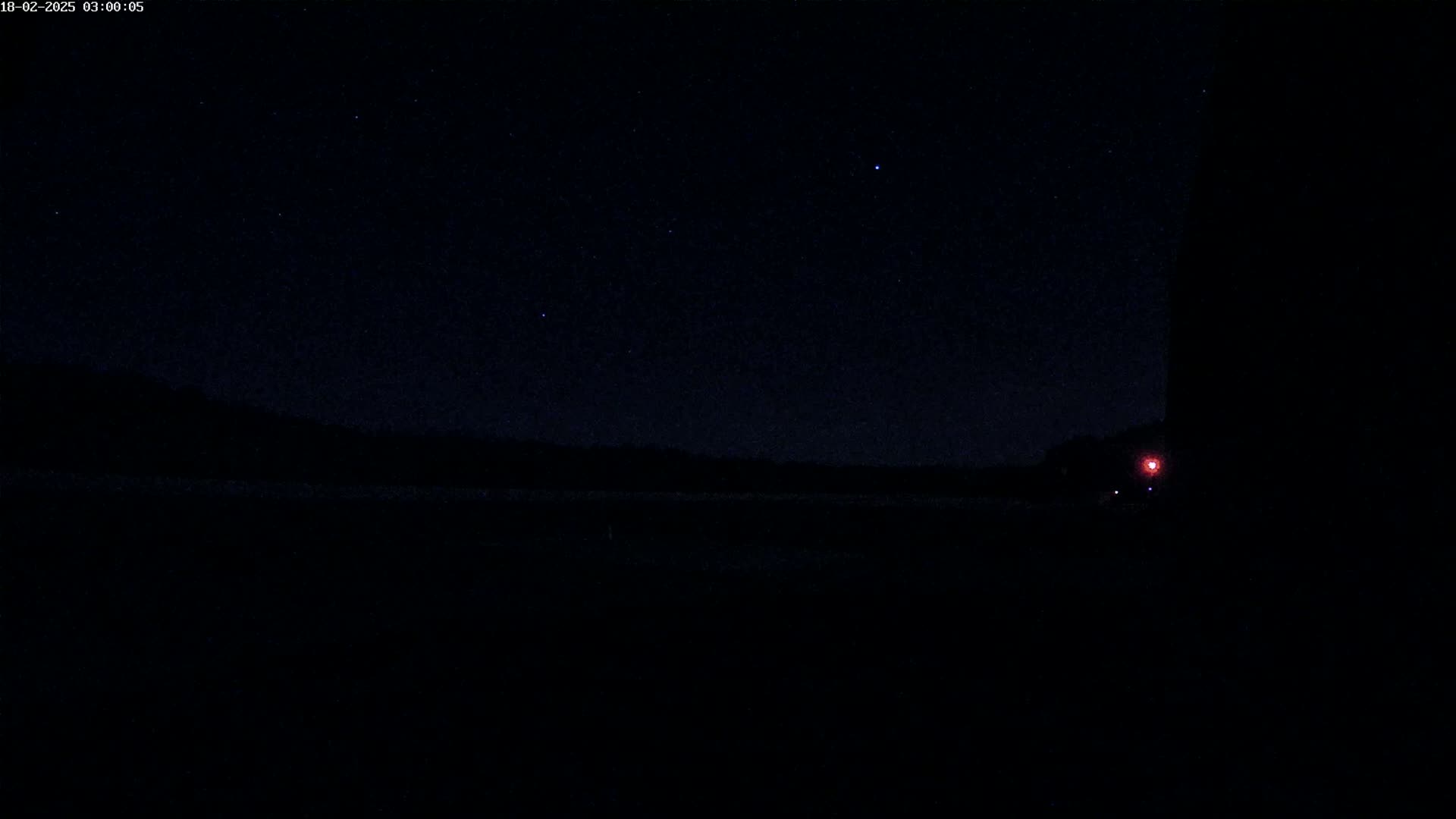 A dark, nighttime landscape with a few visible stars, a distant dark tree line, and a single faint red light in the lower right corner.