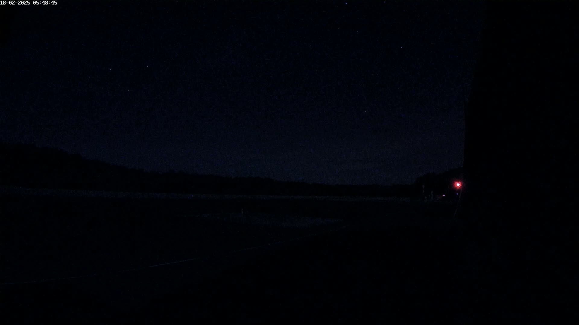 A dark, nighttime scene shows a flat, dark landscape under a starlit sky, with a single red light visible in the distance to the right.