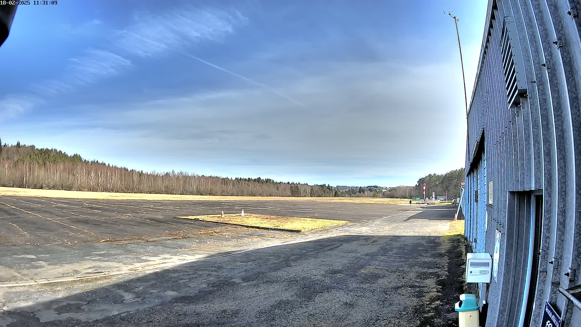 A mostly sunny day reveals a paved airfield bordered by a line of trees, next to a large metal hangar.