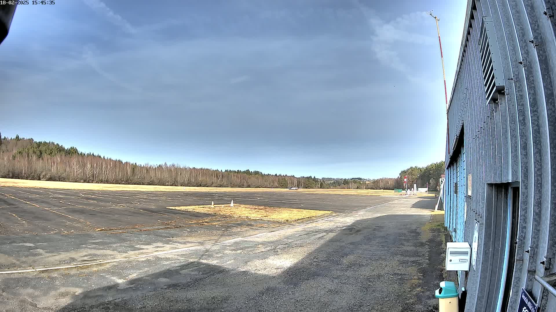 A mostly clear, bright day reveals a paved airfield with a line of trees in the background, next to a corrugated metal building.