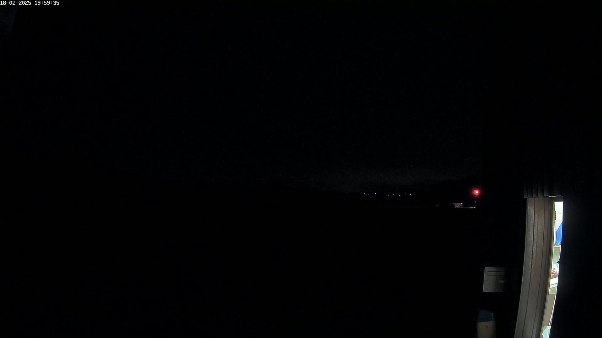 A dark, nighttime scene shows a dimly lit interior doorway and a distant, dark landscape with a few faint lights under a mostly clear sky.