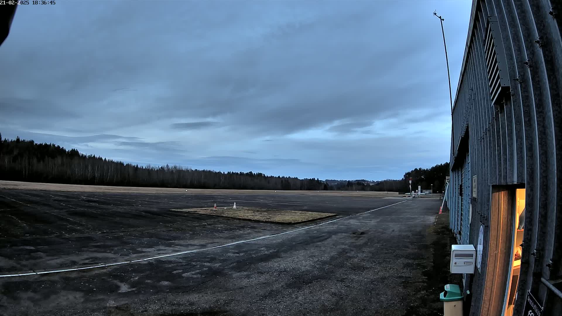 A dark, paved airfield under a cloudy twilight sky is bordered by a line of trees and a hangar with a lit interior.