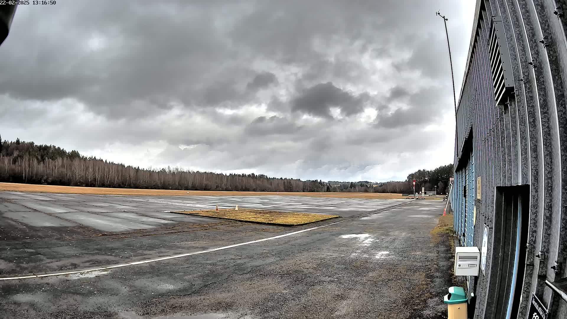 A wet, paved airfield under a cloudy sky is bordered by a line of leafless trees and a corrugated metal building.