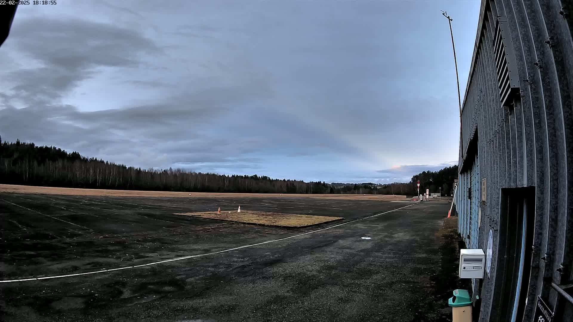 An empty airfield under a cloudy sky is adjacent to a corrugated metal building.
