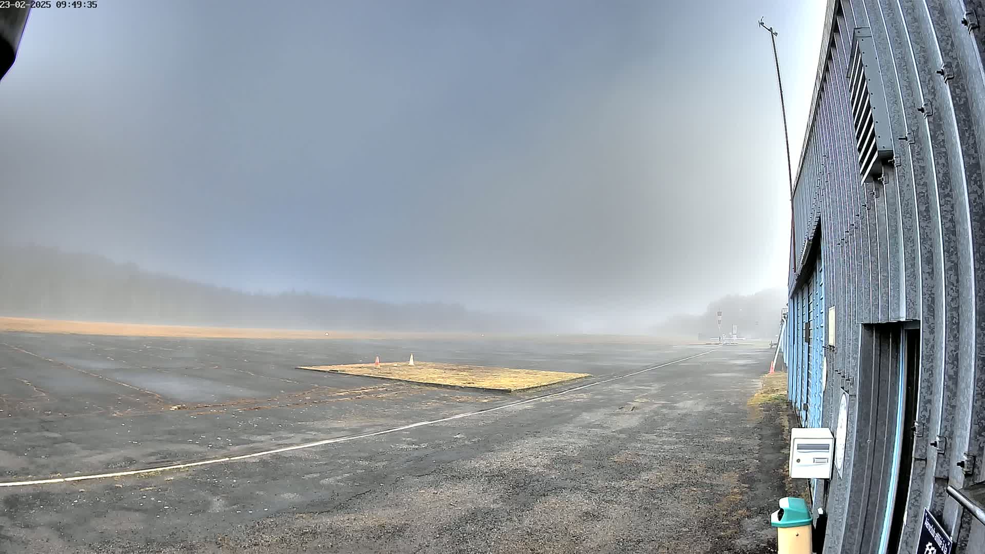 A foggy airfield with a large hangar to the right and a flat, paved area in the foreground.