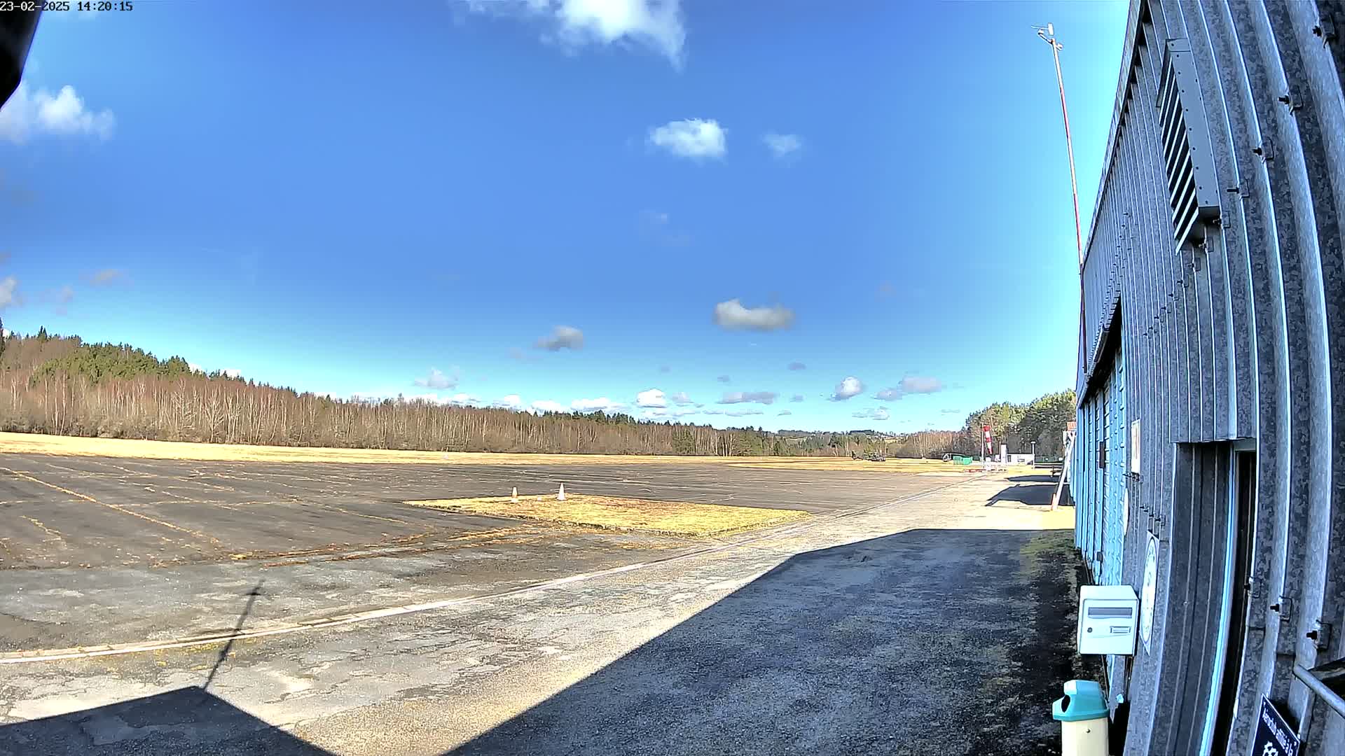 A mostly sunny day reveals a paved airfield bordered by trees, alongside a corrugated metal building.