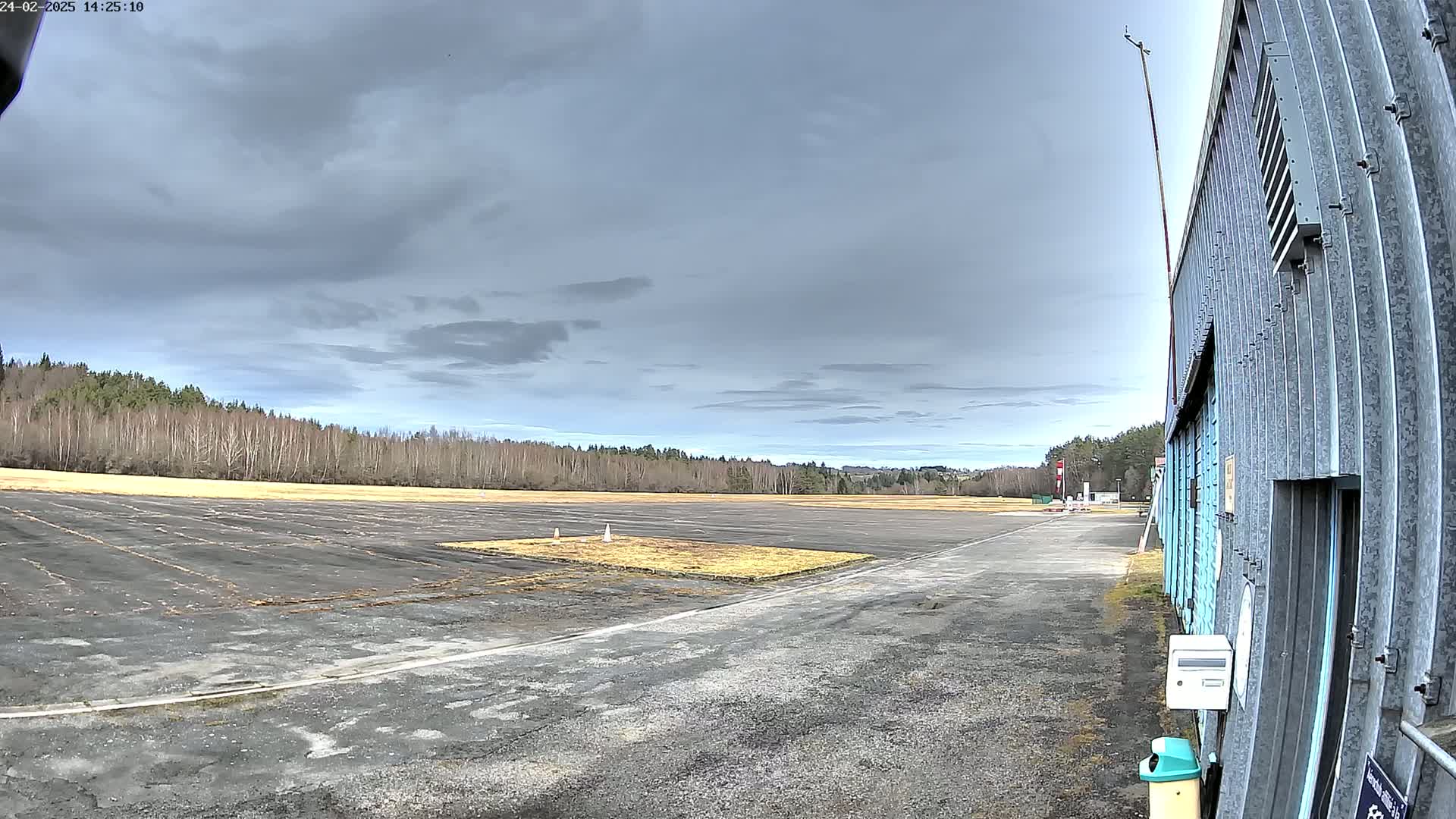 A paved airfield with a grassy area, bordered by a line of trees under a cloudy sky.