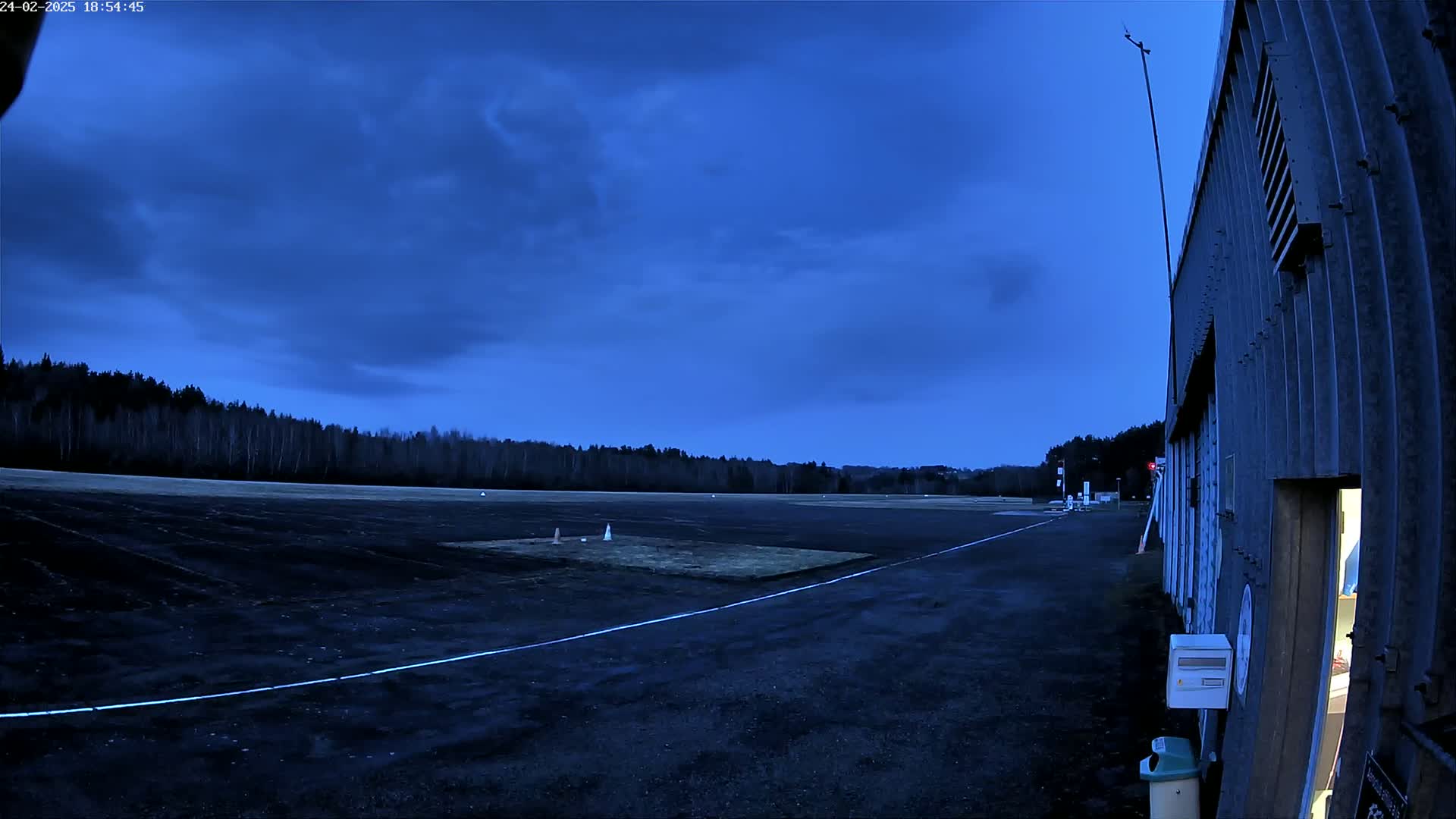 A dark asphalt airfield under a twilight blue sky, bordered by trees and a corrugated metal building with an open door.