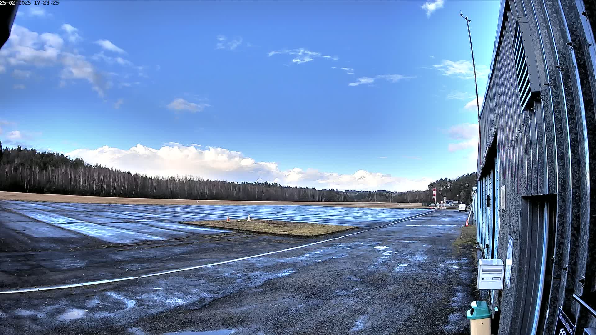 A wet paved area stretches toward a line of trees under a partly cloudy blue sky.