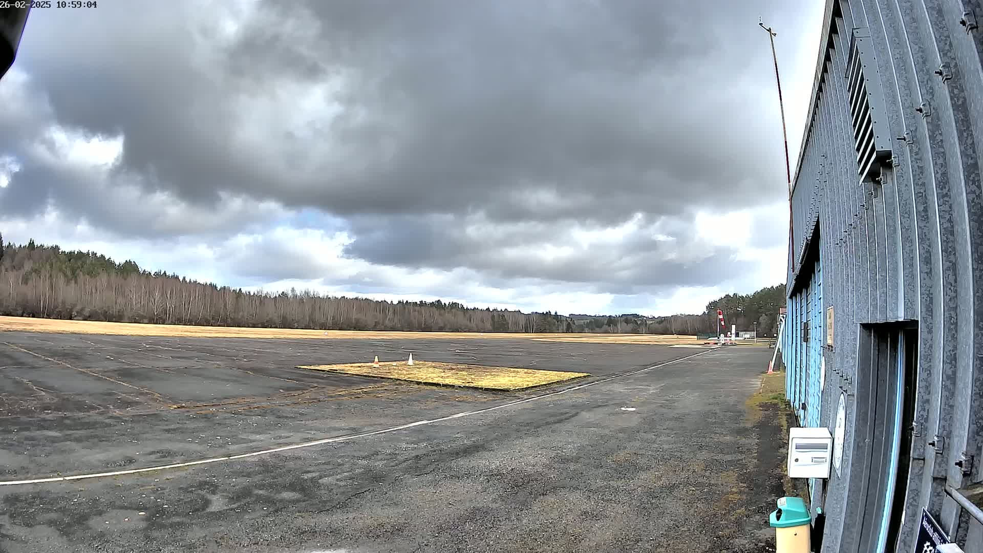 A mostly cloudy sky hangs over a small airfield with a paved runway and a line of trees in the background, next to a corrugated metal building.
