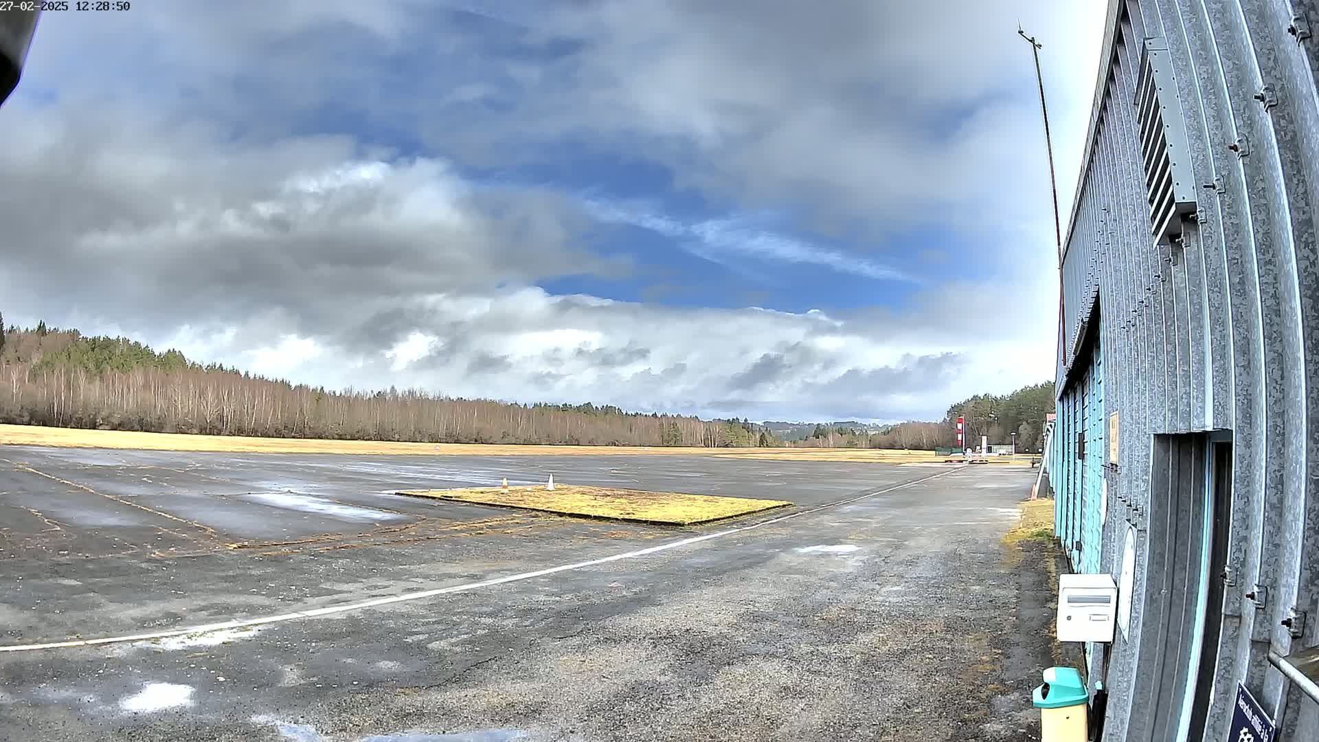 A mostly cloudy sky hangs over a small airfield with a hangar in the foreground, featuring a grassy area in the middle of the tarmac.