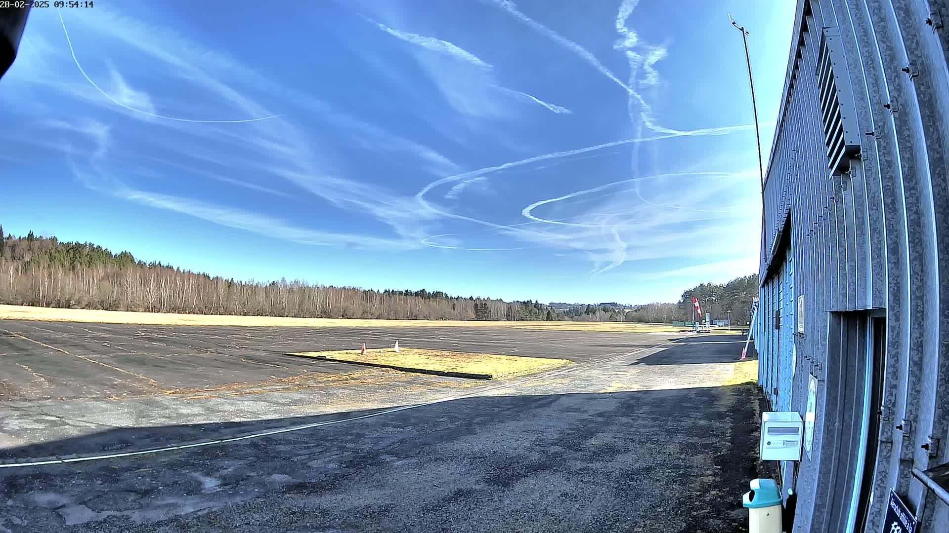 A mostly empty tarmac area next to a long metal building under a bright blue sky with contrails.