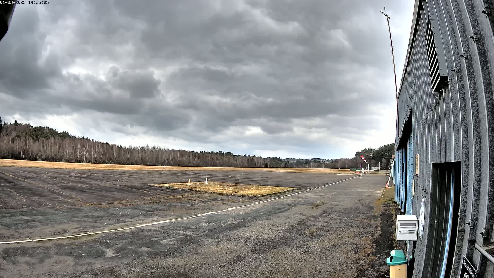 A dark, overcast sky hangs over a small airfield with a paved runway and a long, corrugated metal building.