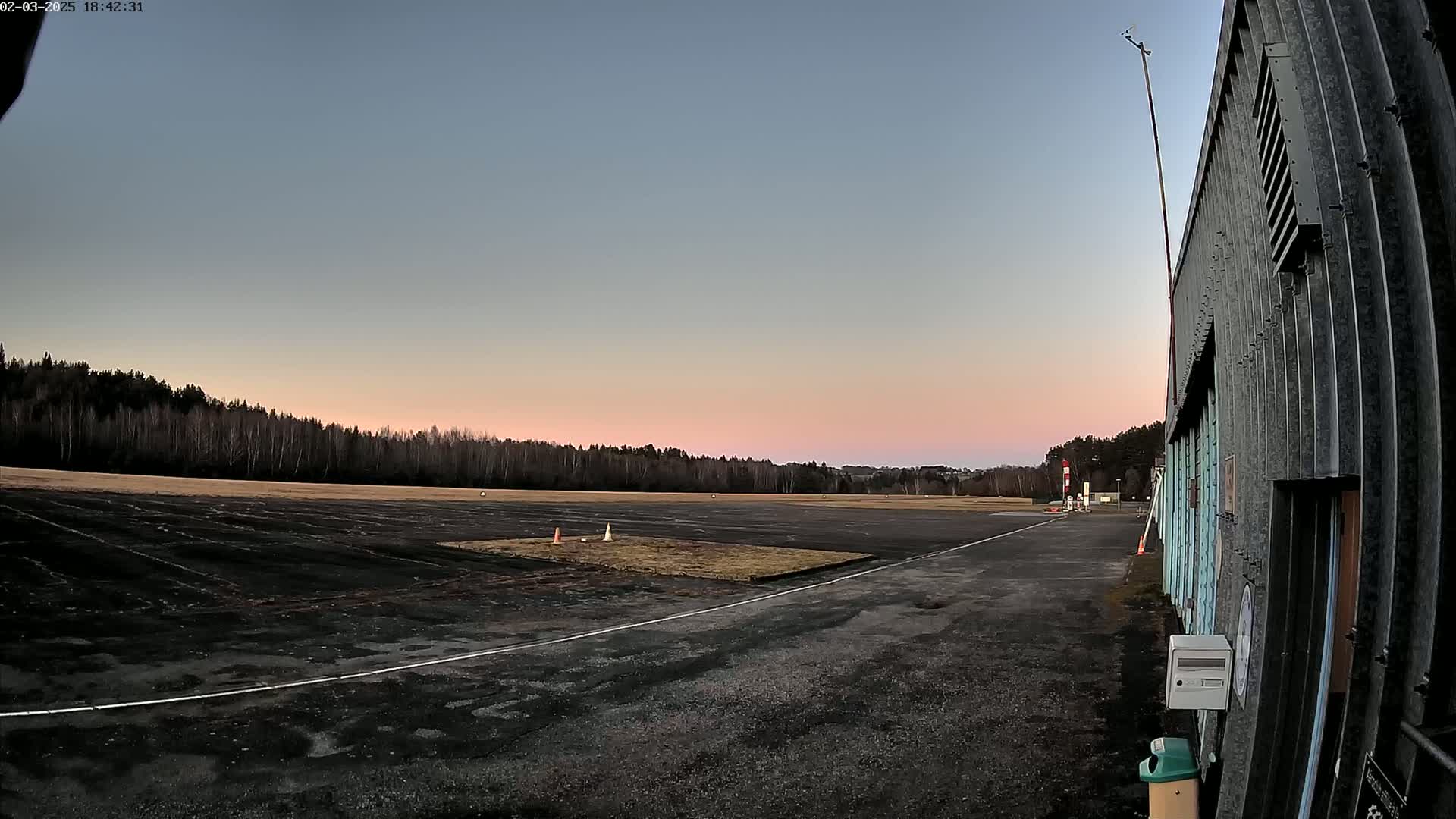 A paved airfield with a line painted on it sits at the edge of a line of bare trees under a twilight sky.