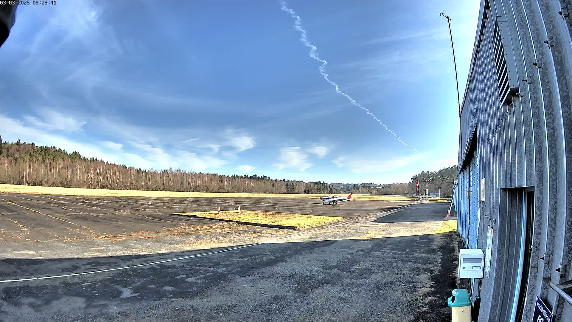A small airplane sits on a tarmac next to a hangar under a mostly sunny sky with a contrail in the distance.