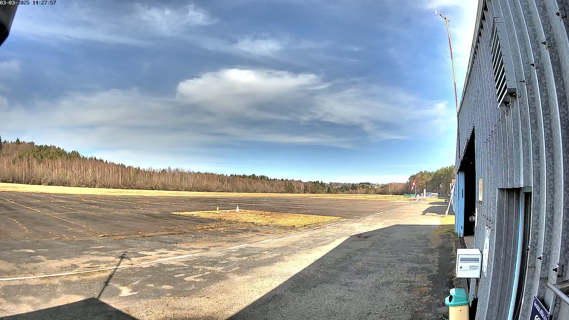 A mostly sunny day reveals a paved airfield bordered by a line of trees and a corrugated metal building.