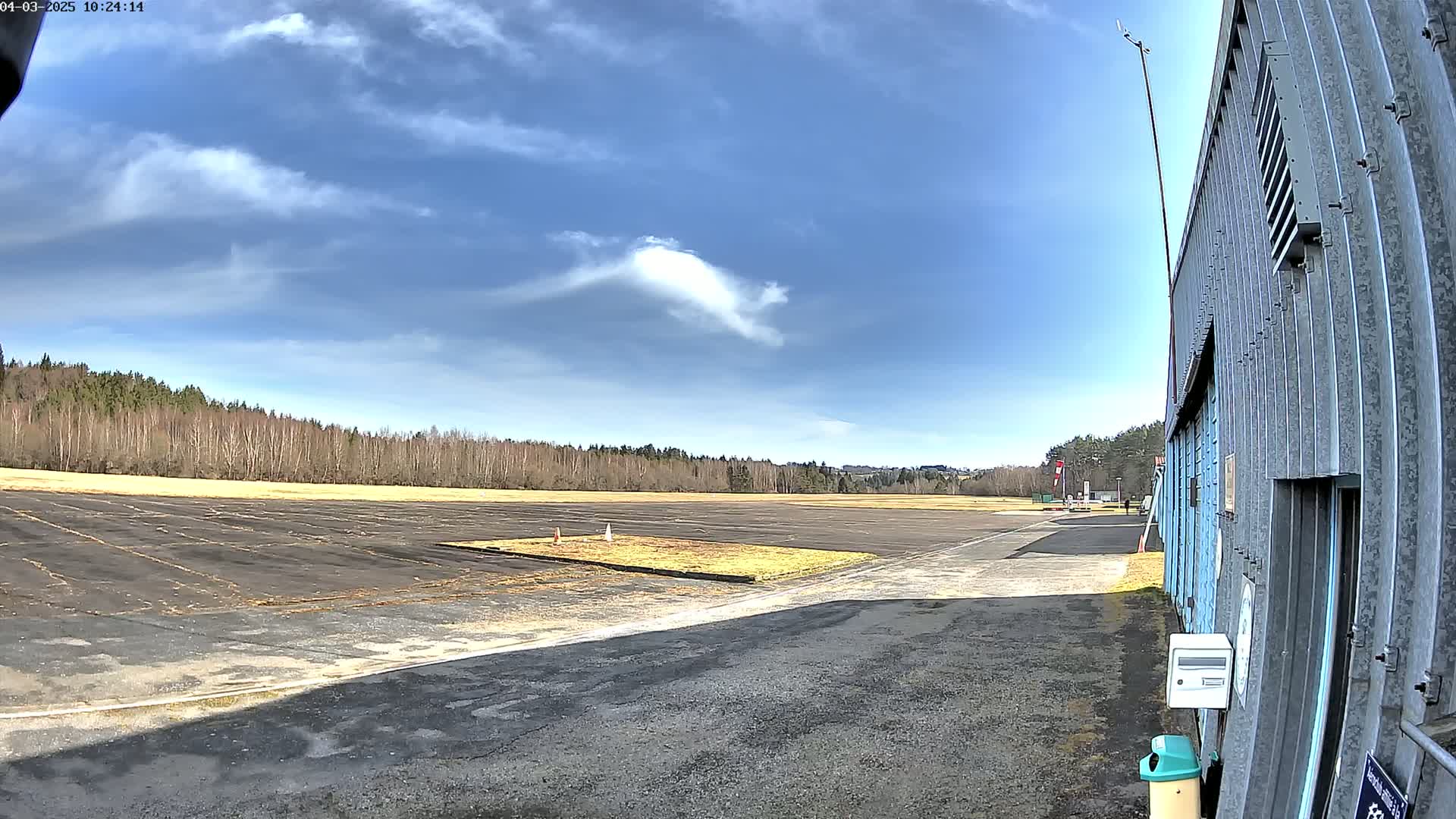 A mostly sunny day reveals a paved airfield bordered by trees, with a portion of a hangar visible to the right.