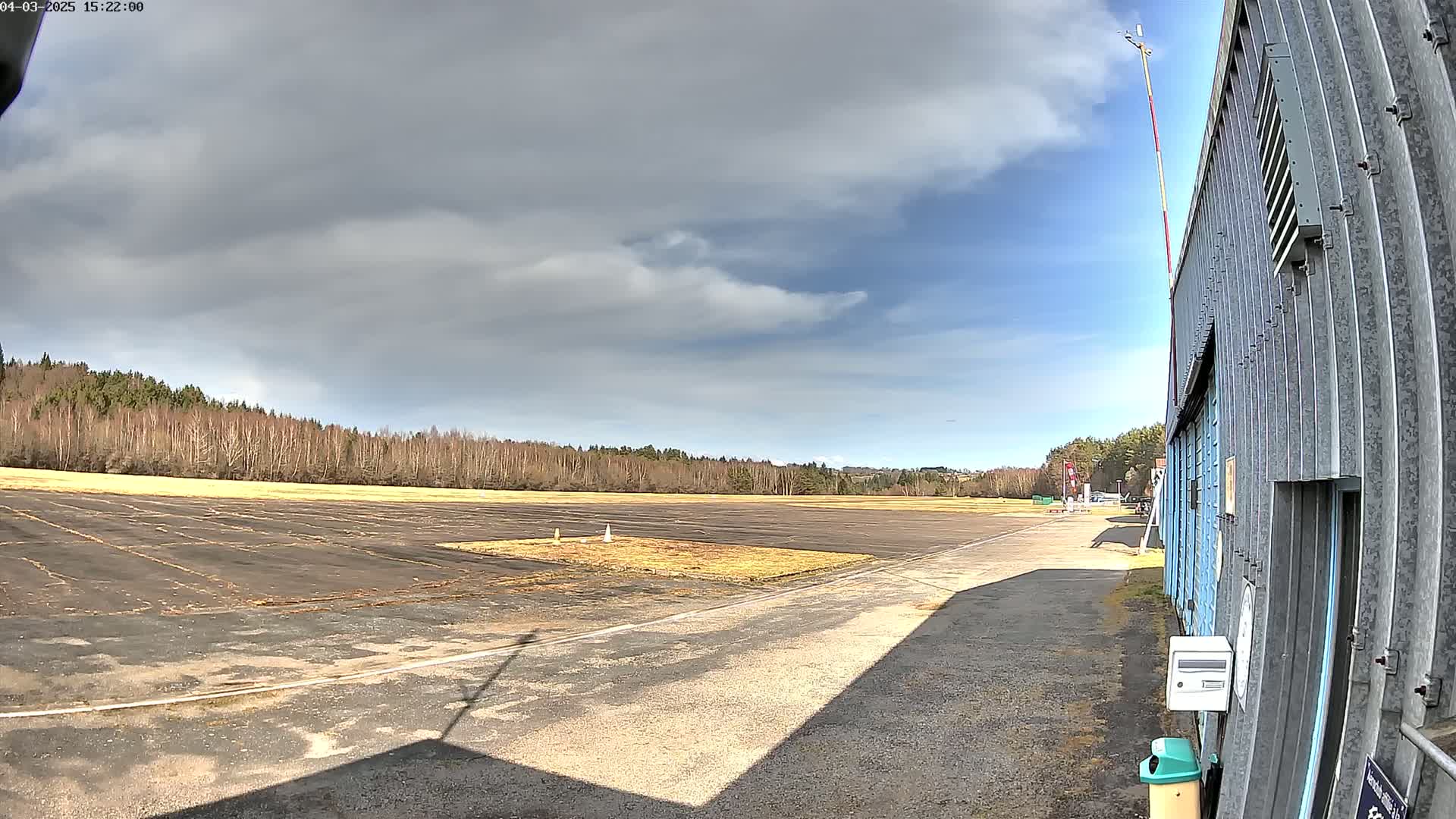 A mostly sunny day with partly cloudy skies shows a tarmac airfield next to a corrugated metal building, bordered by a line of leafless trees.